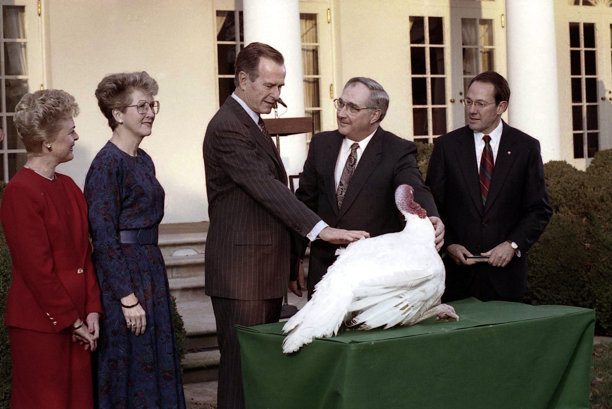 Five adults stand outdoors around a table with a large white turkey. The man in the center, wearing a suit, is touching the turkey. The group looks happy and engaged. The setting appears formal and official.
