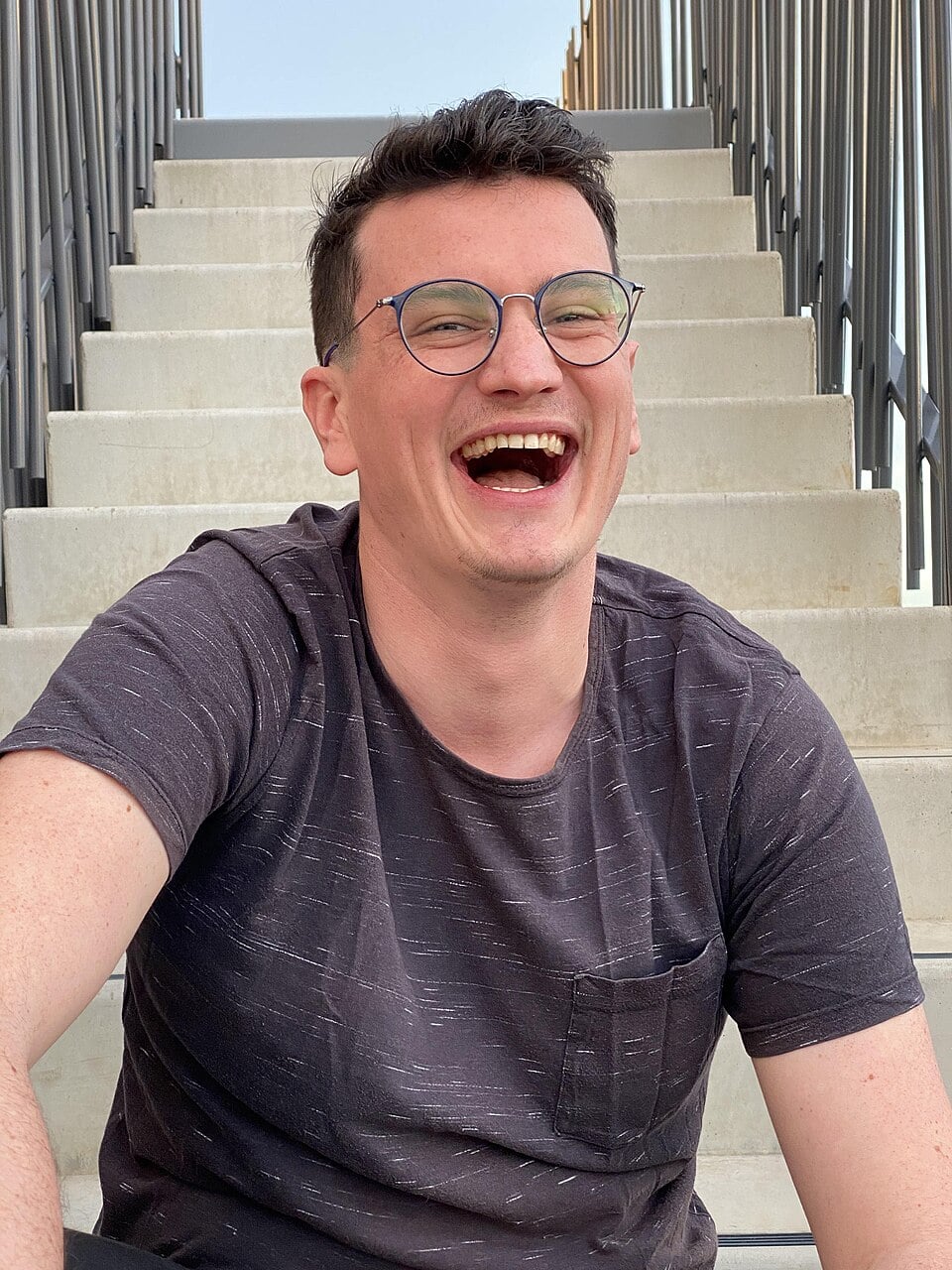 A smiling person with short dark hair and glasses, wearing a dark t-shirt, sits on outdoor concrete steps, laughing joyfully. Metal railings line both sides of the steps.