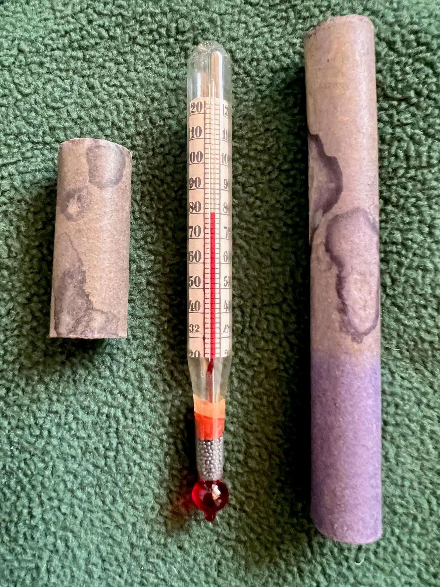 A glass thermometer with a red liquid center is shown between two parts of a cylindrical, purple and gray paper case, all placed on a green textured surface.