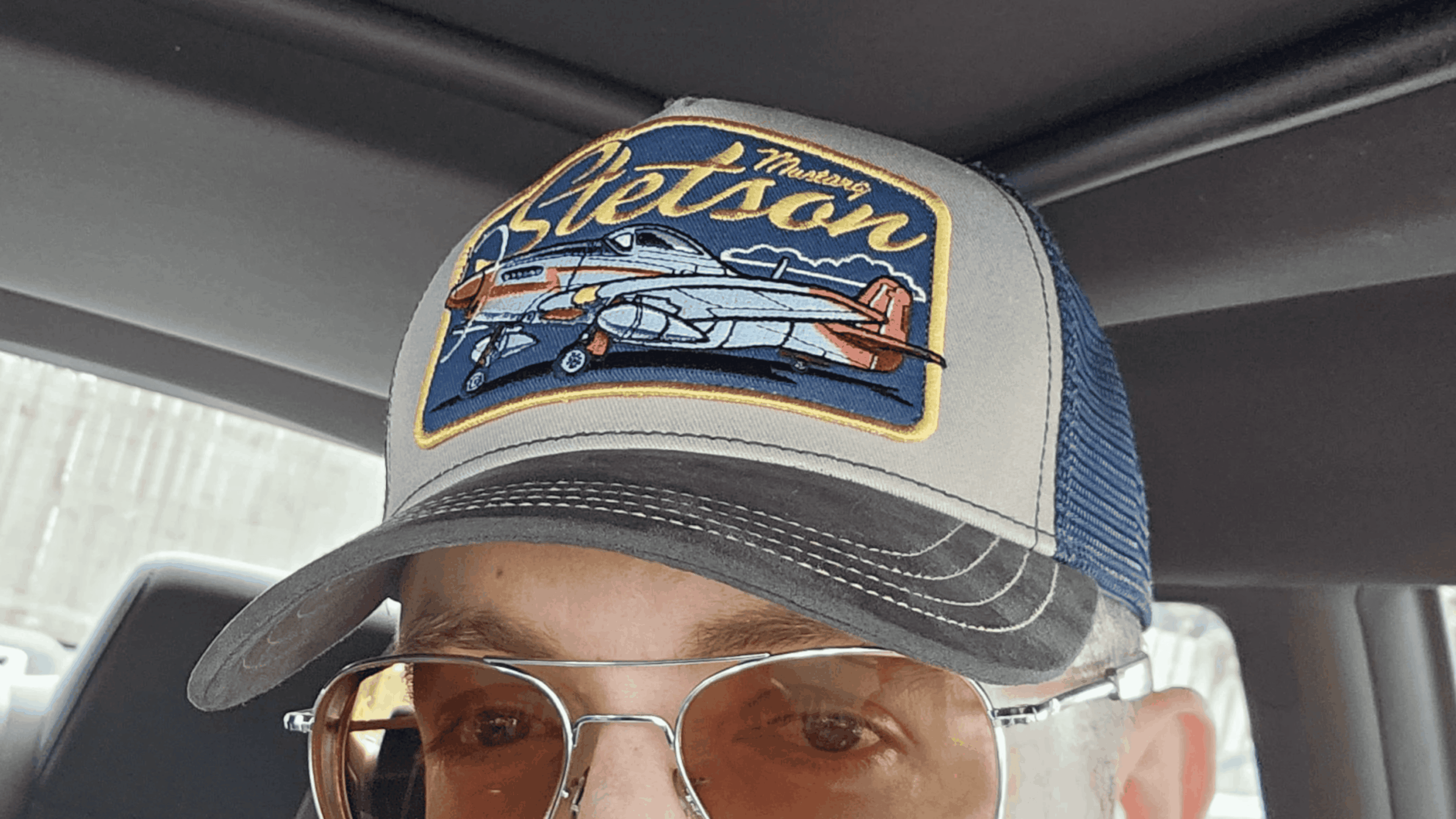A person wearing sunglasses and a trucker hat with a Stetson logo and an illustrated airplane patch, sitting inside a car.