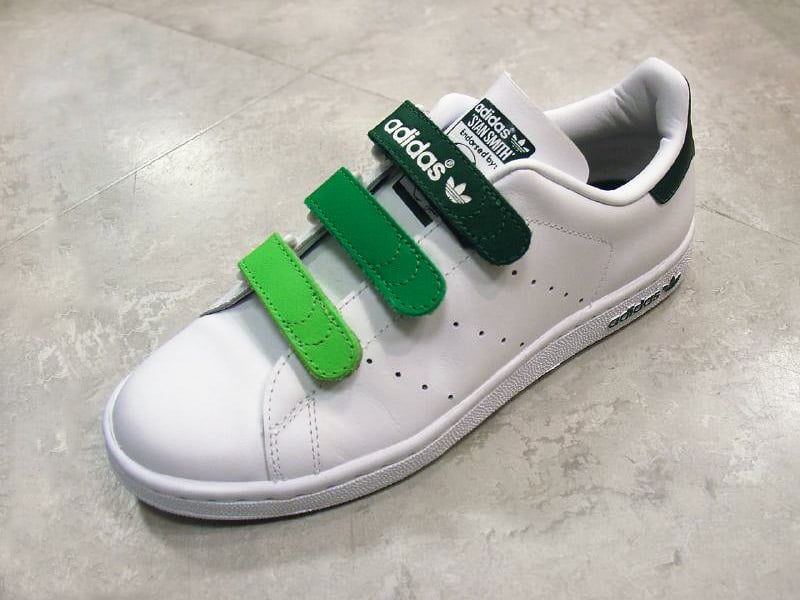 A white Adidas sneaker with three Velcro straps in varying shades of green sits on a light gray tile floor. The shoe features dark green details on the heel and branding on the tongue and side.
