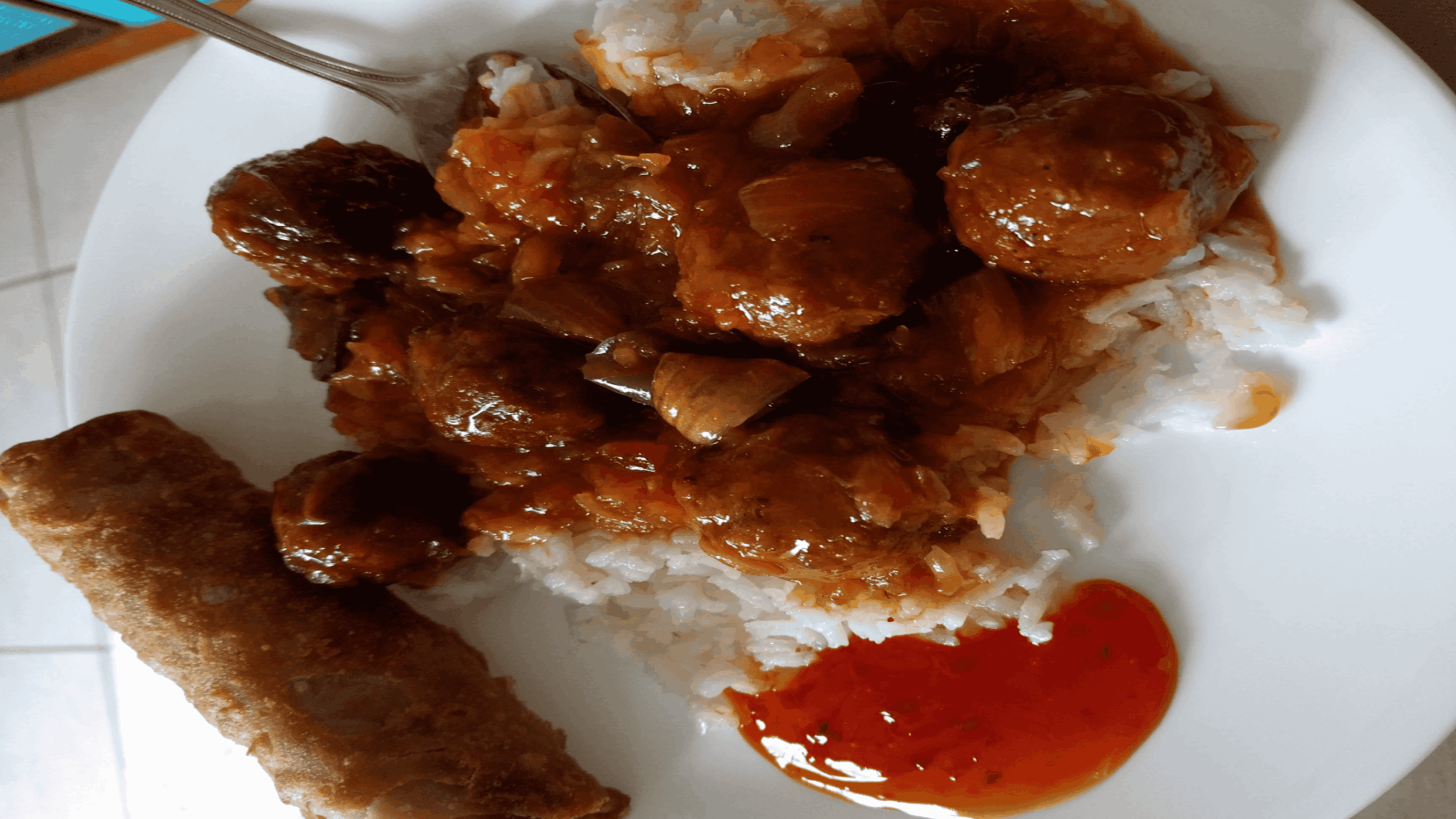 A plate of white rice topped with meatballs in brown sauce, served with a spring roll and a dollop of red sauce on the side.
