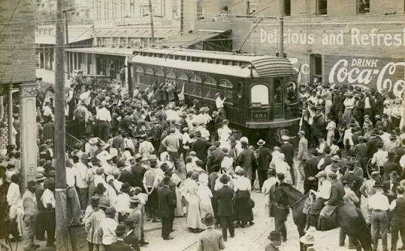 Large crowd gathers around a streetcar on a busy city street; vintage clothing suggests early 20th century. Brick buildings with Coca-Cola ads are visible in the background.