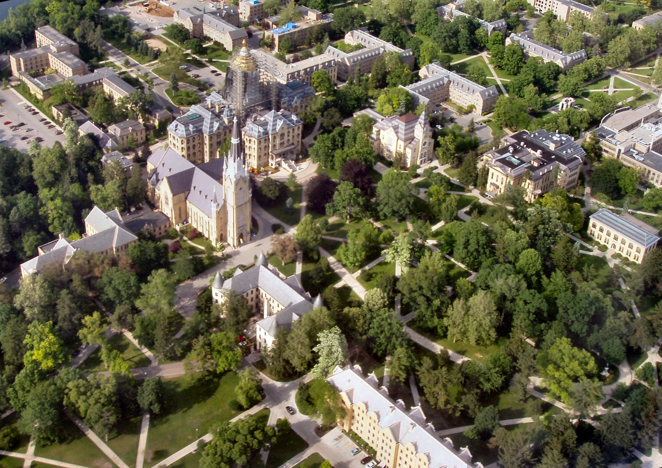 Aerial view of a university campus with historic buildings, tree-lined paths, and green lawns. Several large structures, including a church with a tall spire, are surrounded by lush landscaping.