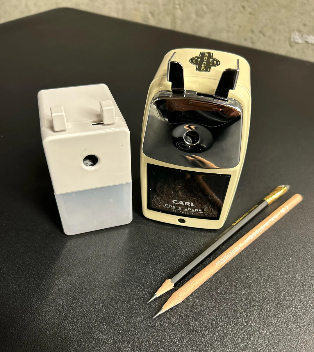 A manual pencil sharpener, its detachable shaving container, and two sharpened pencils with erasers are placed on a black surface.