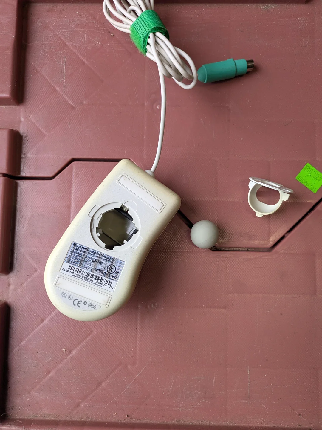 A disassembled old computer mouse with the bottom cover and trackball removed, resting on a textured brown surface. The green PS/2 connector and white cable are visible.