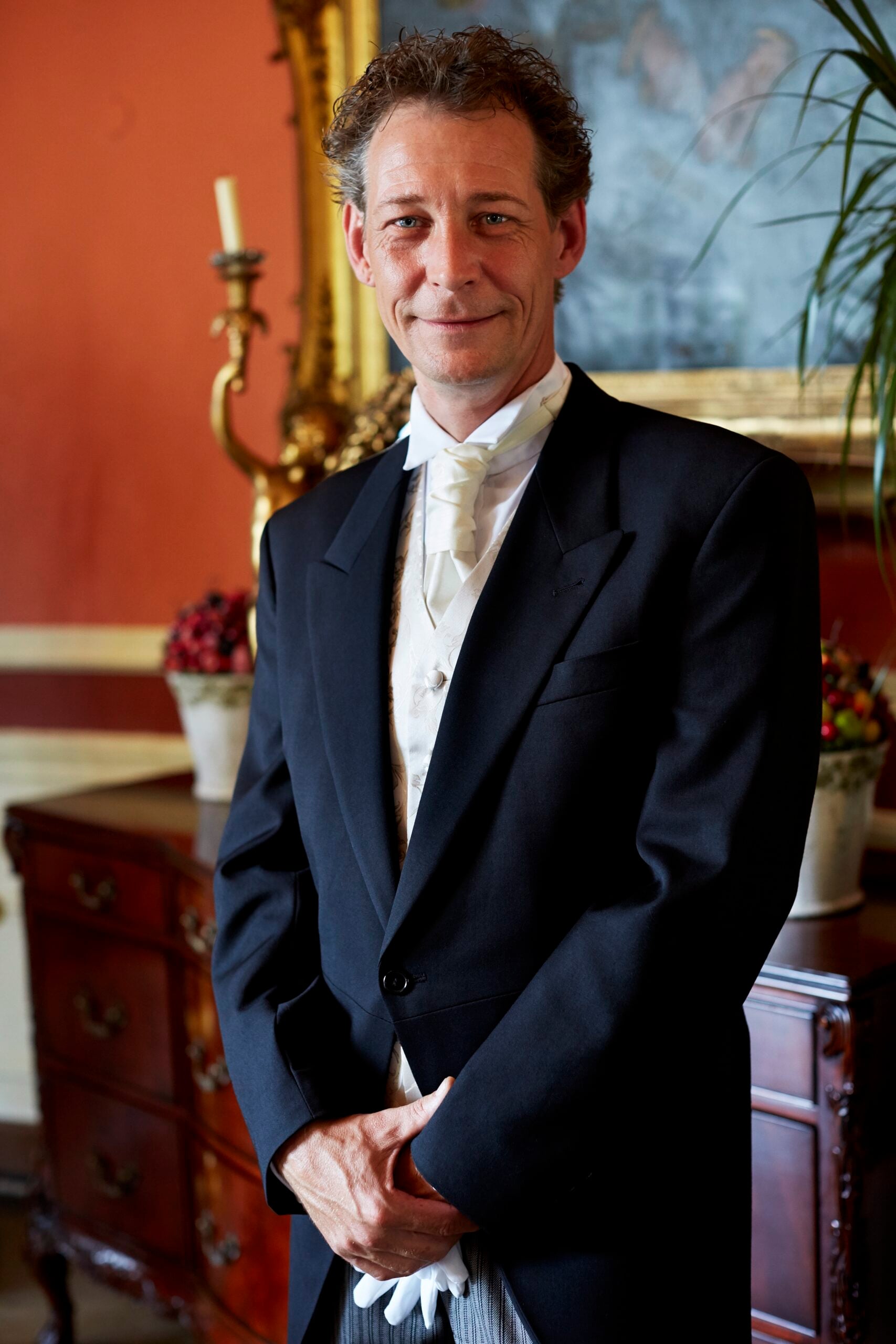 A man in formal attire, wearing a black tailcoat, white bow tie, and gloves, stands indoors in an elegant room with ornate decor, a dresser, and a framed painting behind him.