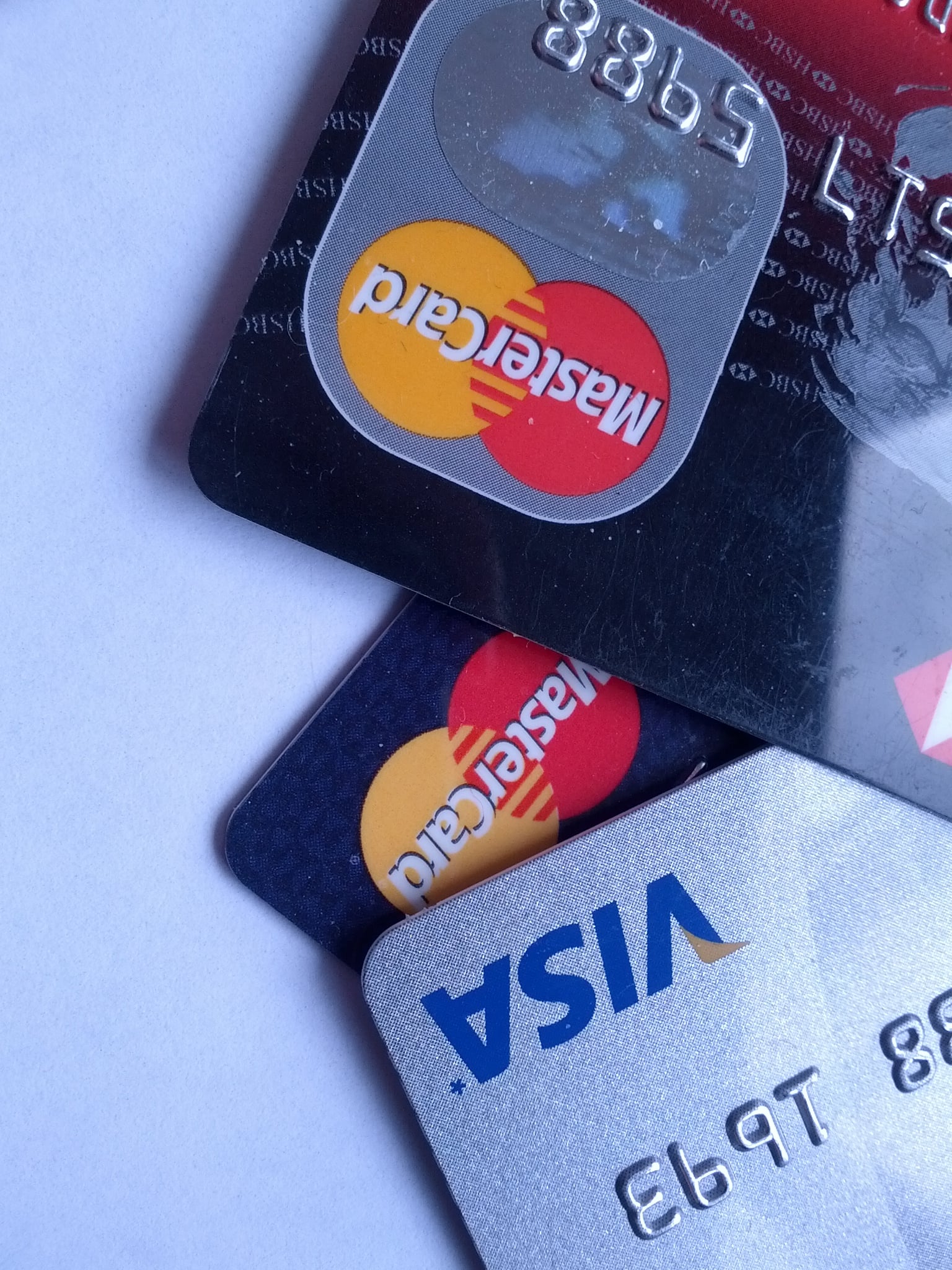 Three overlapping credit cards: two Mastercards and one Visa, all placed on a white surface. The cards are positioned at an angle, showing parts of the chip, logos, and embossed numbers.