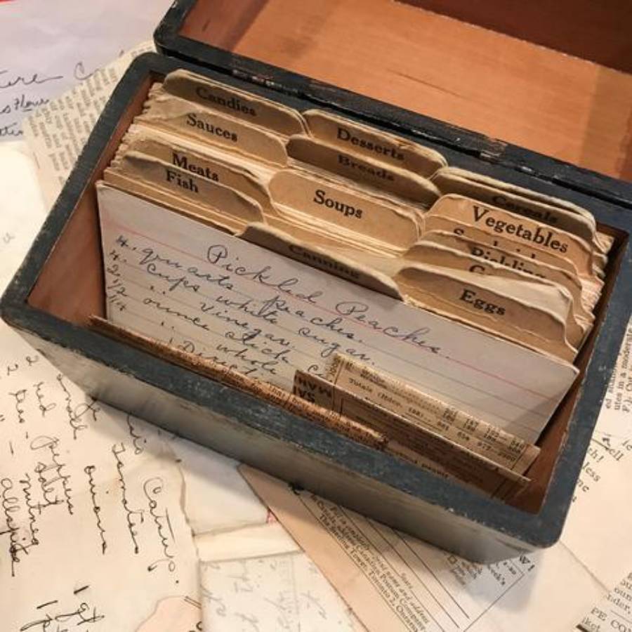 An open recipe box with labeled dividers and handwritten recipe cards inside. Papers and handwritten notes are scattered around the box on the table.