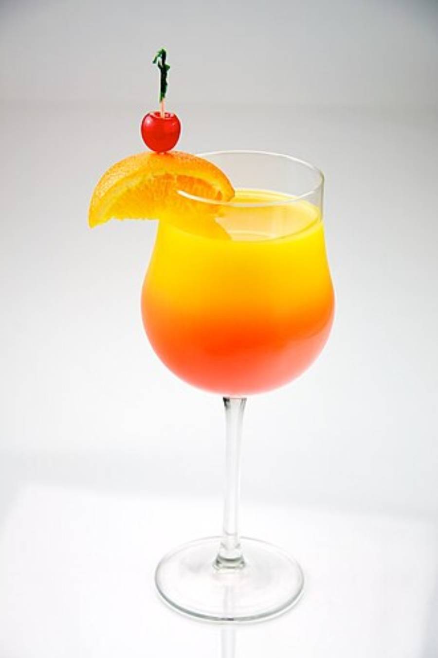 A vibrant orange and red layered cocktail in a stemmed glass, garnished with an orange slice and a cherry on a toothpick, set against a plain white background.