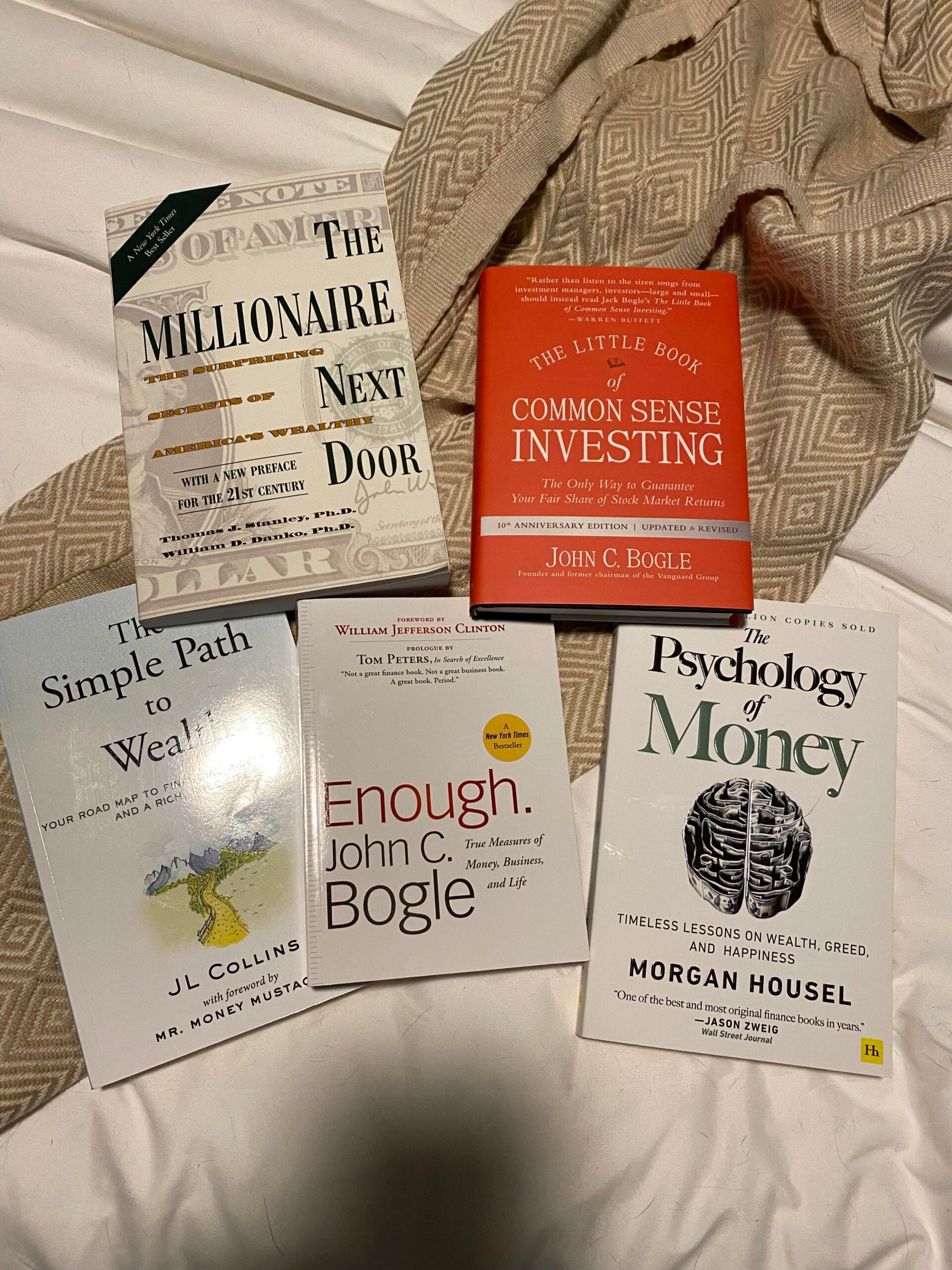 Five personal finance books, including “The Millionaire Next Door,” “The Little Book of Common Sense Investing,” “The Simple Path to Wealth,” “Enough,” and “The Psychology of Money,” arranged on a white blanket.
