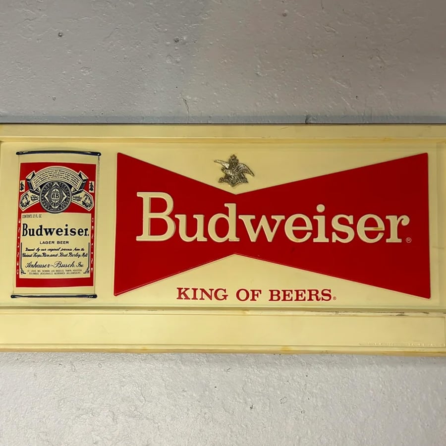 A vintage Budweiser sign features a can on the left and a large red bowtie logo with “Budweiser” and “King of Beers” text on a cream background.