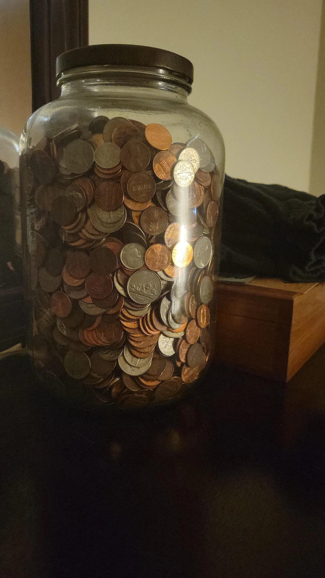 A large glass jar filled with assorted coins sits on a dark surface next to a wooden box, with light reflecting off the glass.