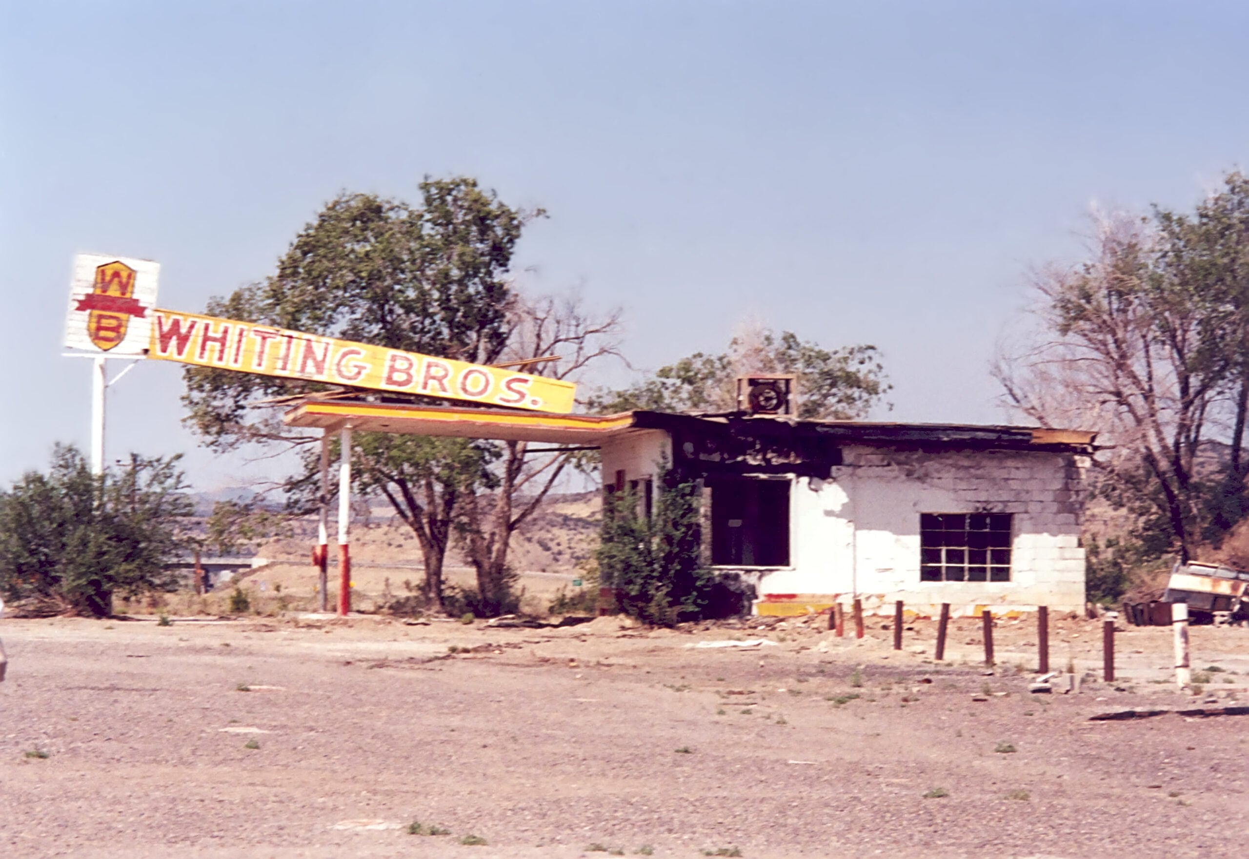 An abandoned gas station with a faded “Whiting Bros.” sign sits in a dry, empty lot, surrounded by trees and brush under a clear blue sky.