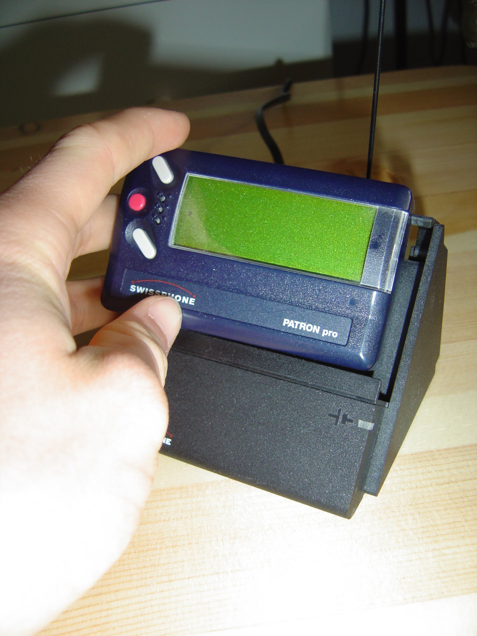 A hand holds a blue Swissphone Patron Pro pager, placing it into a black charging dock on a wooden surface.