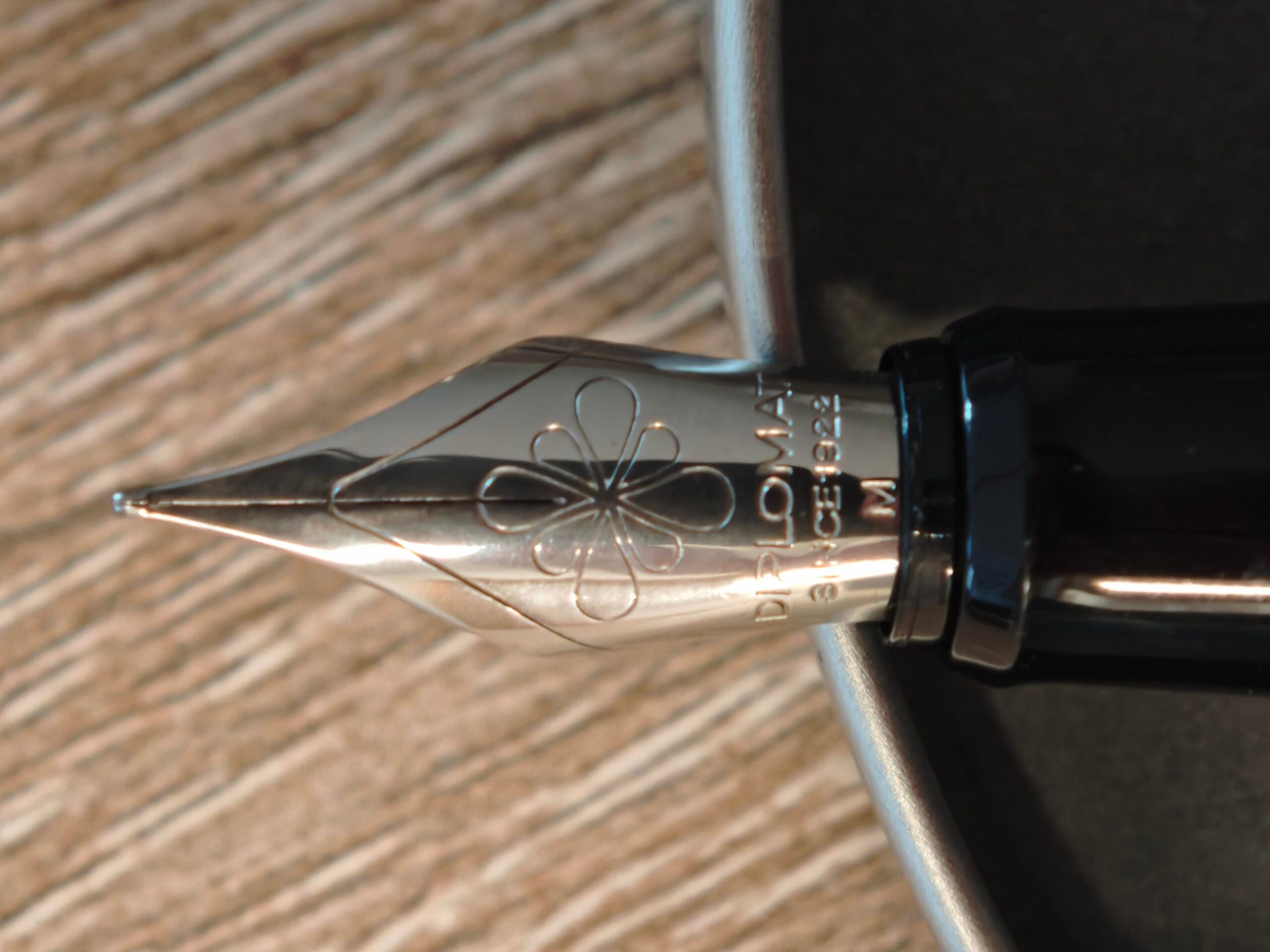 Close-up of a silver fountain pen nib resting on the edge of a gray dish, with a wooden surface underneath. The nib has an engraved flower design and the word “DIPLOMAT” is partially visible.