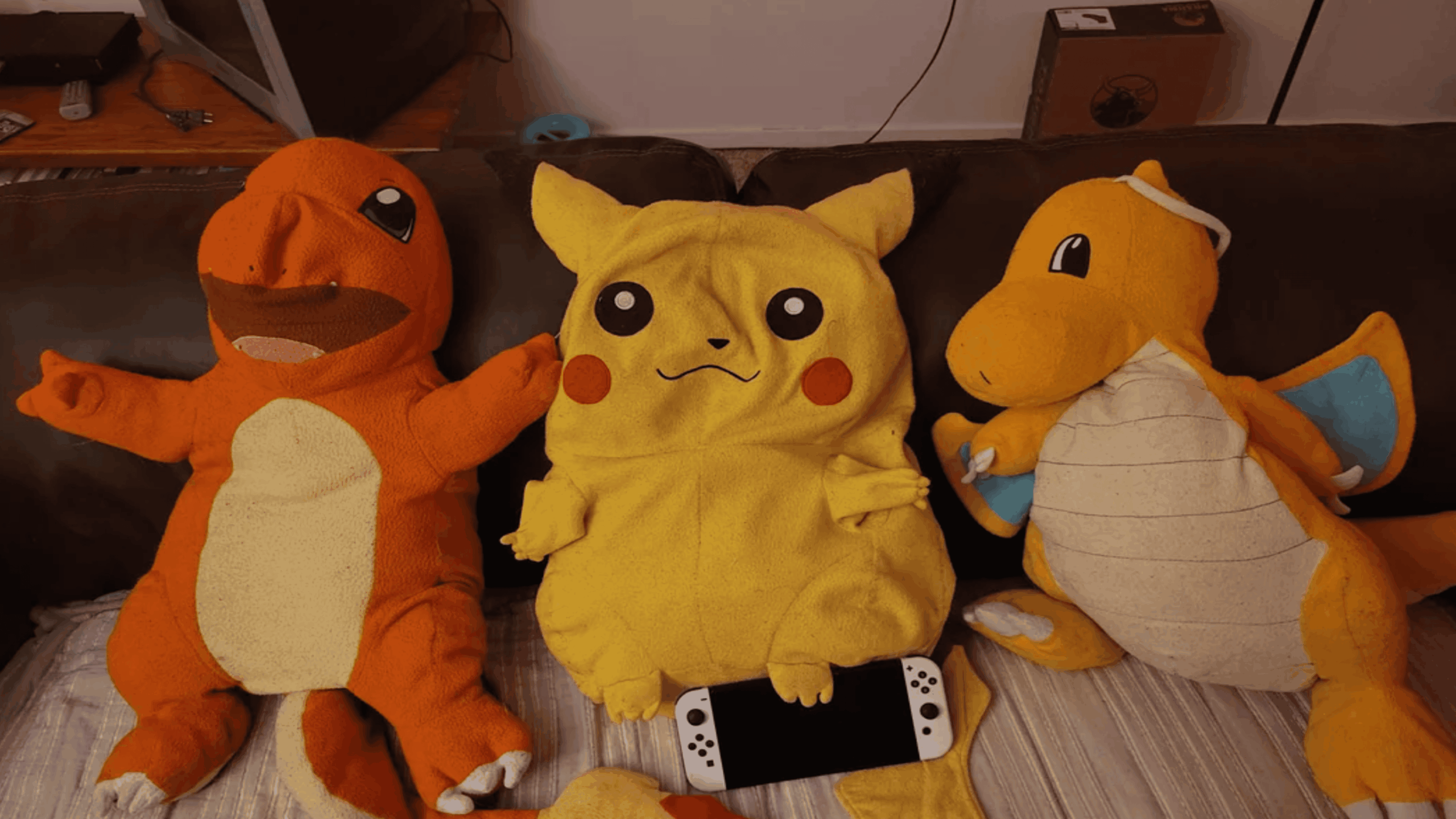Three plush toys—Charmander, Pikachu, and Dragonite—sit on a couch. Pikachu holds a Nintendo Switch Lite in its lap. A cardboard box and other household items are in the background.
