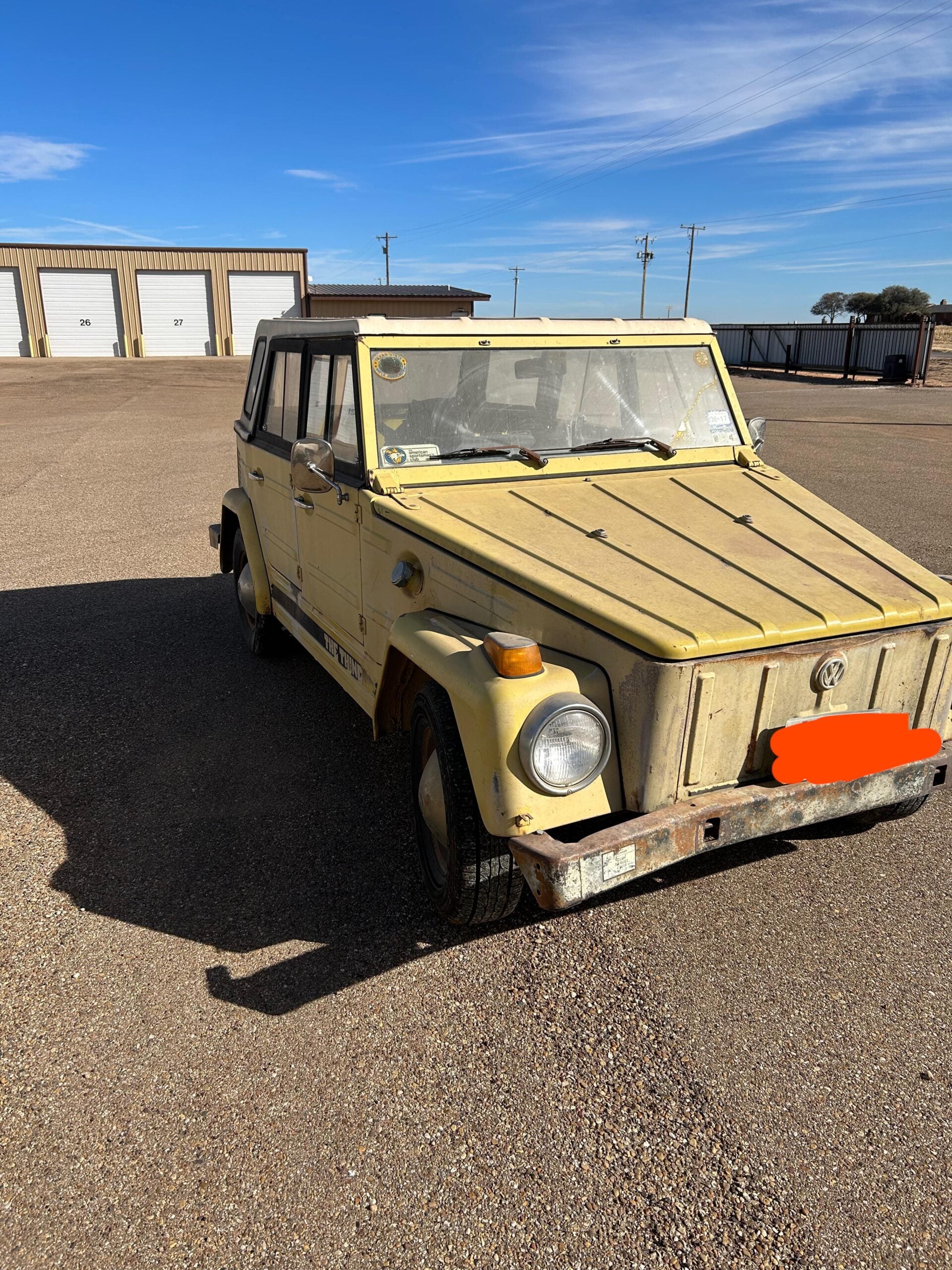 A weathered, yellow vintage Volkswagen car parked on a gravel lot with warehouse buildings in the background. The car has rust and visible wear.