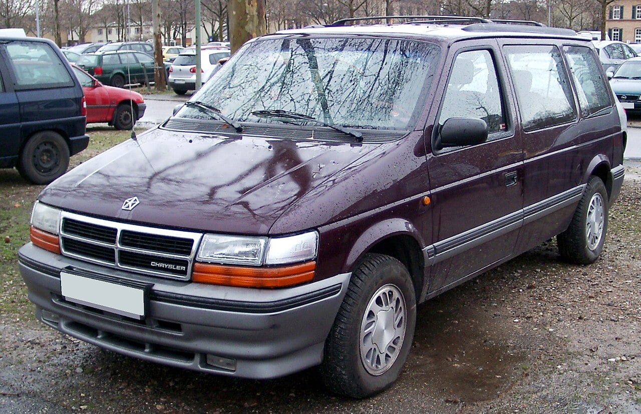 A maroon Chrysler minivan from the 1990s is parked in a lot among other vehicles. The van has a boxy shape, silver wheels, and water droplets on its surface.