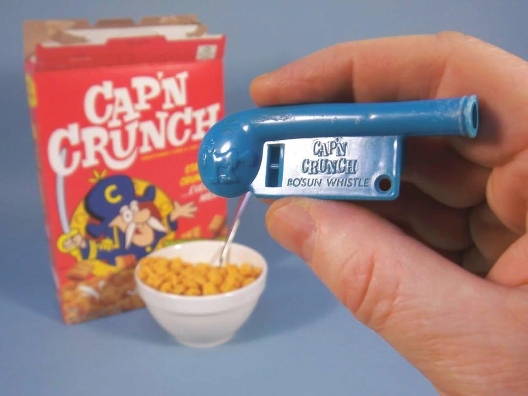 A hand holds a blue Cap’n Crunch Bosun Whistle. In the background, a Cap’n Crunch cereal box and a bowl of cereal with a spoon are visible.