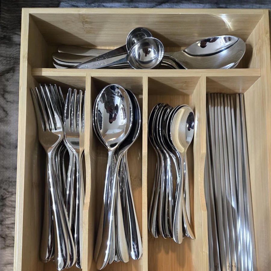 A wooden drawer organizer holding metal forks, spoons, teaspoons, knives, and measuring spoons arranged neatly in separate compartments.