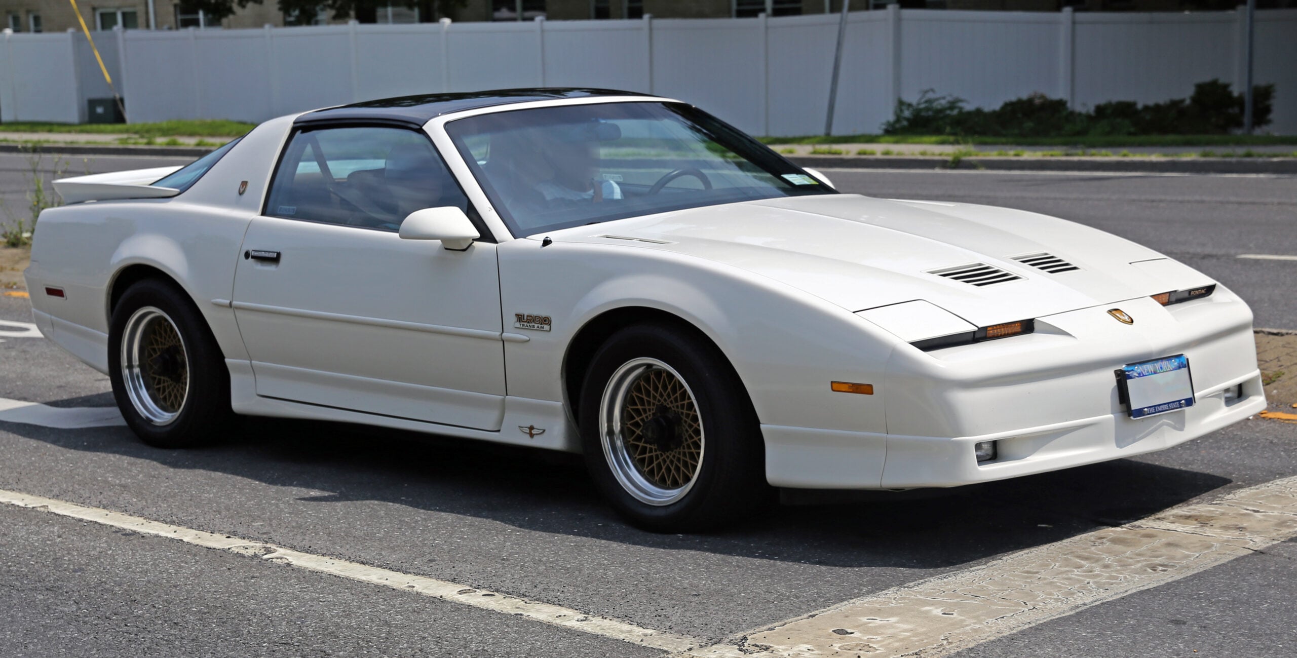 A white sports car with pop-up headlights and T-top roof is parked on the street near a curb, featuring gold-spoke wheels and a sleek, 1980s-style design.