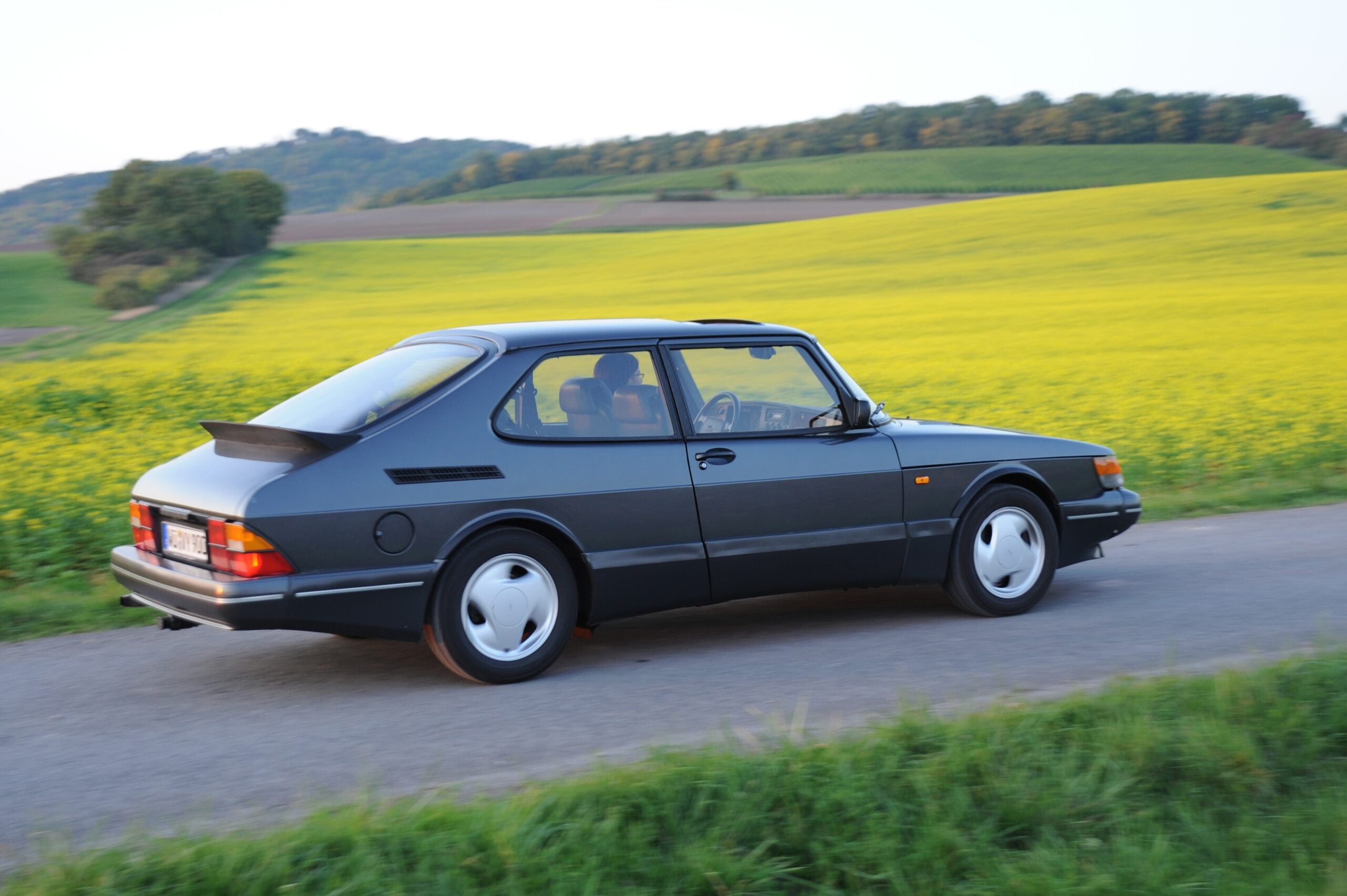 A dark gray vintage Saab car drives on a rural road, bordered by green grass and yellow fields, with gentle hills and trees in the background under a clear sky.