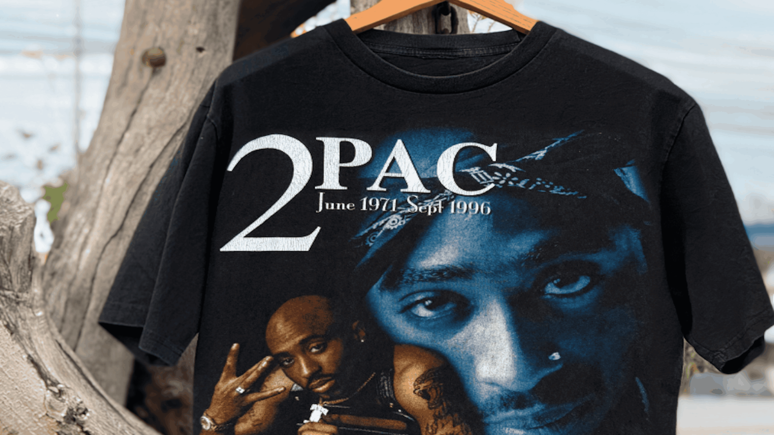 A black T-shirt featuring large text “2PAC June 1971 – Sept 1996” and two images of Tupac Shakur’s face, one with a bandana and the other with his hand showing a peace sign.