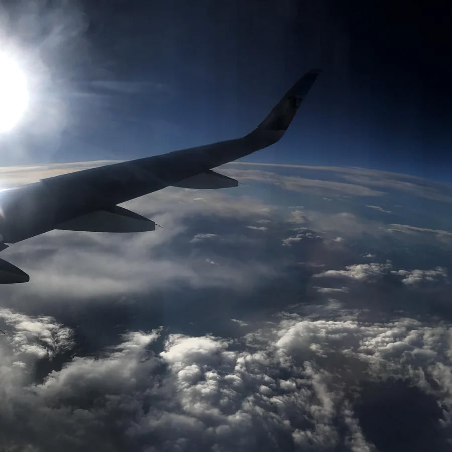View from an airplane window showing a wing, clouds below, and a bright sun partially obscured, with a dark blue sky above.