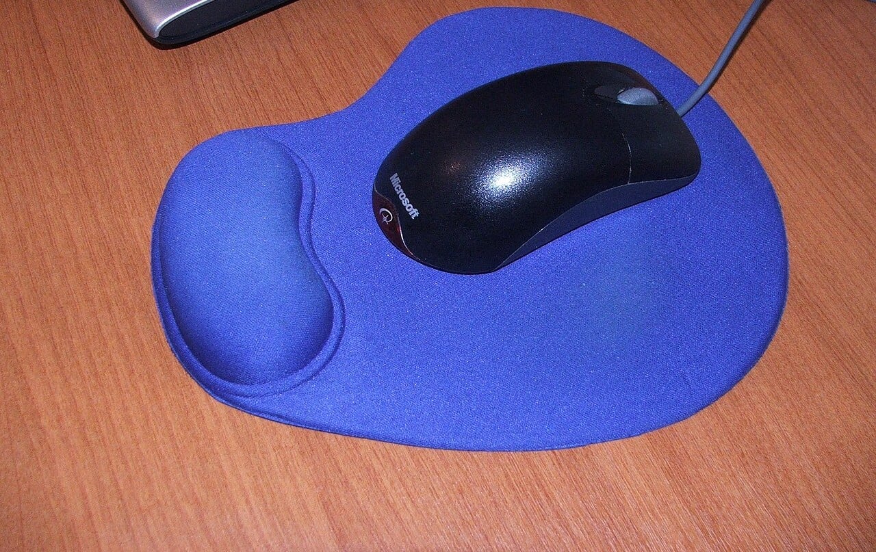 A black computer mouse on a blue mouse pad with a built-in wrist rest, placed on a wooden desk.