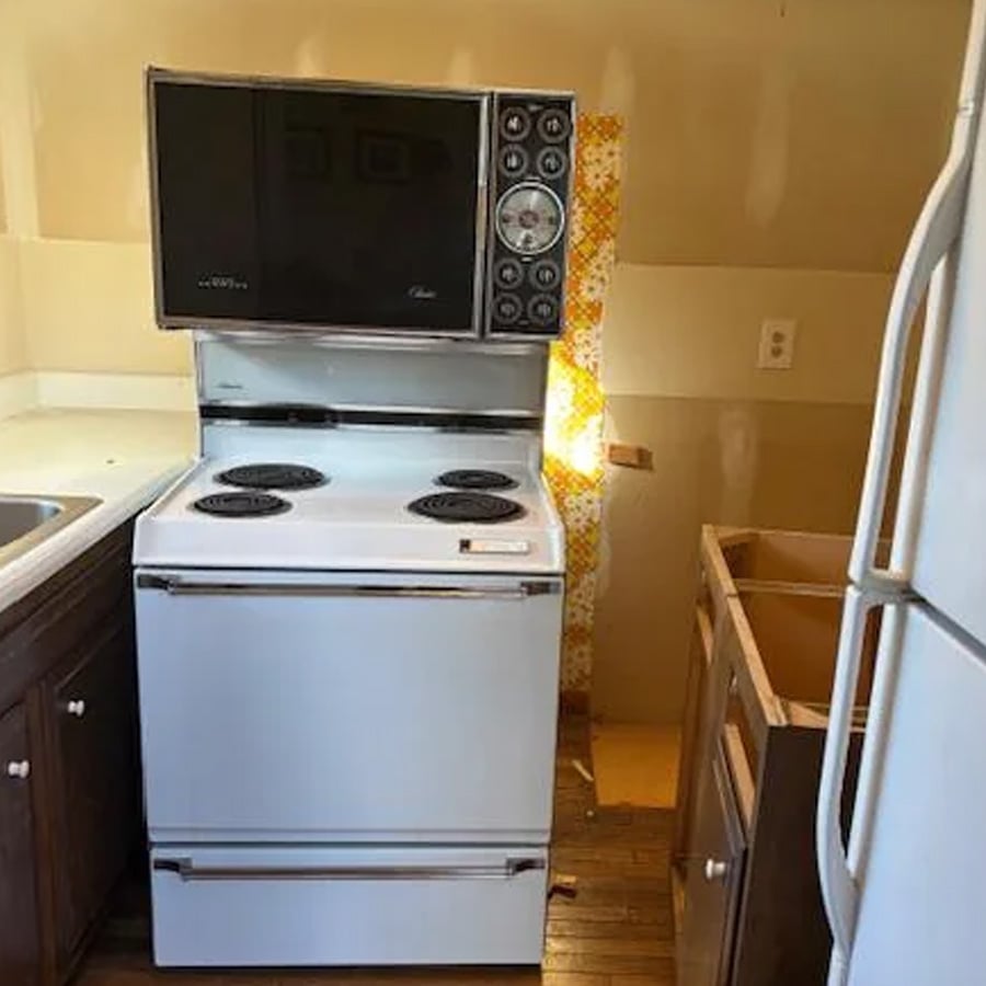 A vintage white stove with electric burners and an oven, topped by a microwave with many dials, sits in an unfinished kitchen. Nearby are a refrigerator, empty cabinet, and a strip of yellow floral wallpaper.