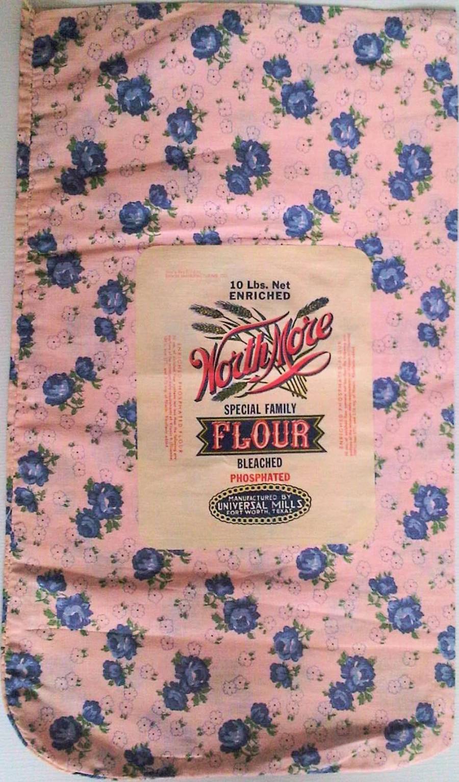 A vintage cloth flour sack with a pink background and blue flowers, featuring a label that reads “Worth More Special Family Flour” in red and black text. The sack is slightly worn but still vibrant.