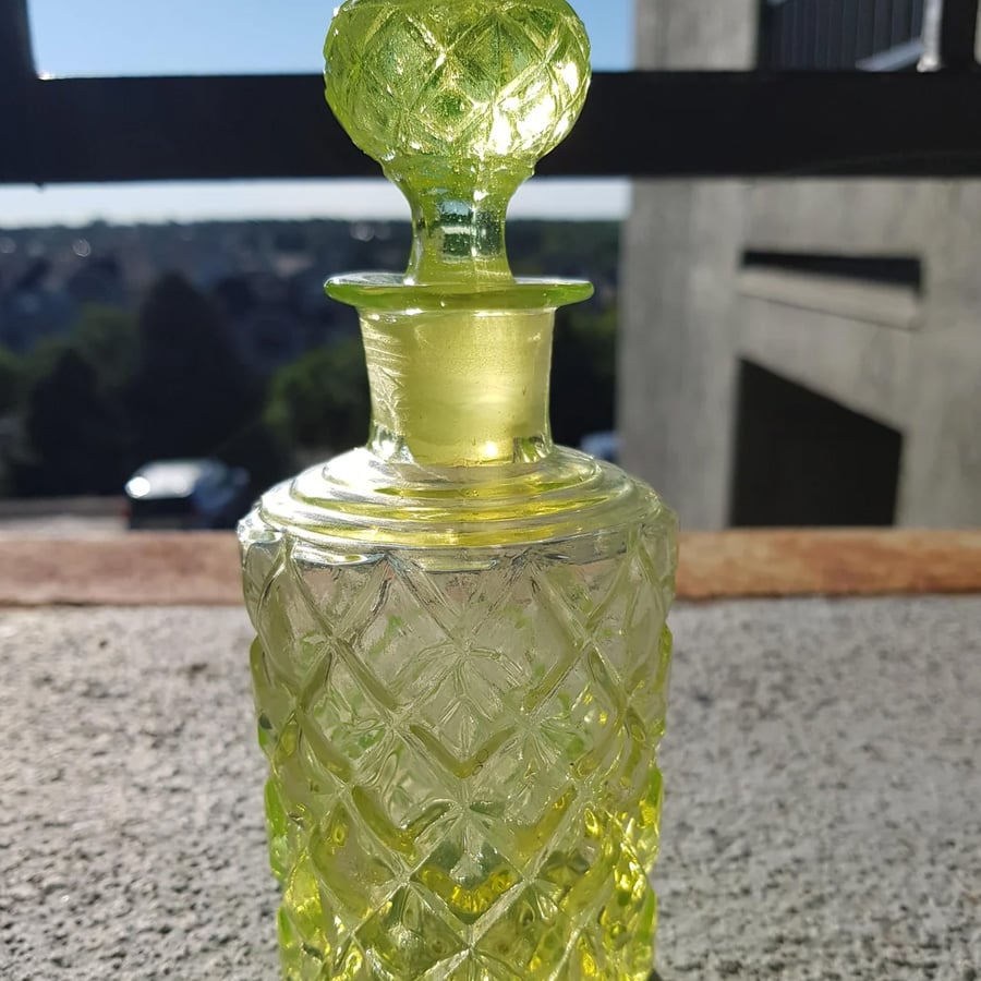 A vintage yellow-green glass decanter with a textured diamond pattern sits on a sunlit outdoor surface, with a blurred background of trees, cars, and buildings.