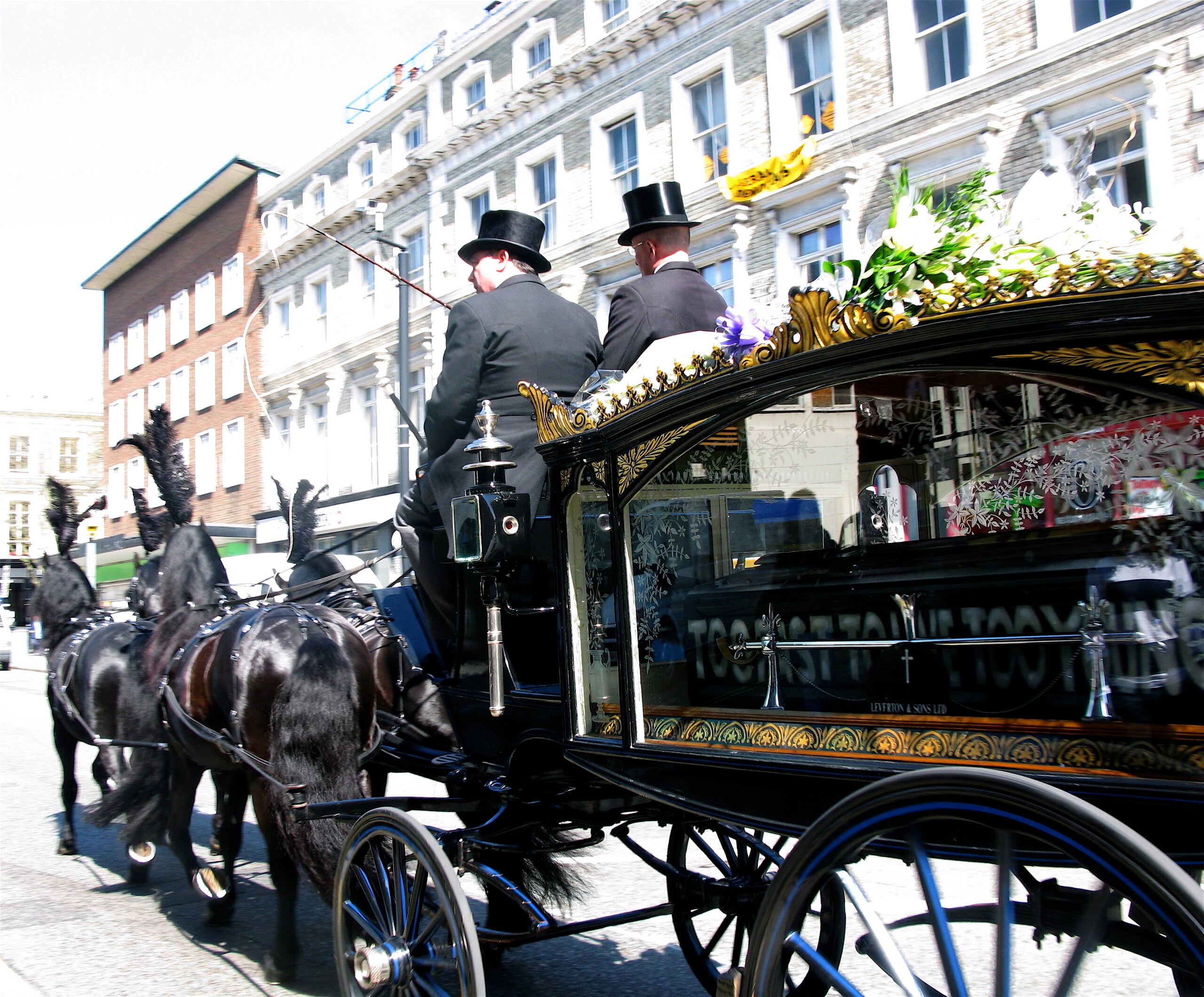 A black horse-drawn hearse with two men in top hats transports a casket adorned with flowers through a city street lined with buildings.