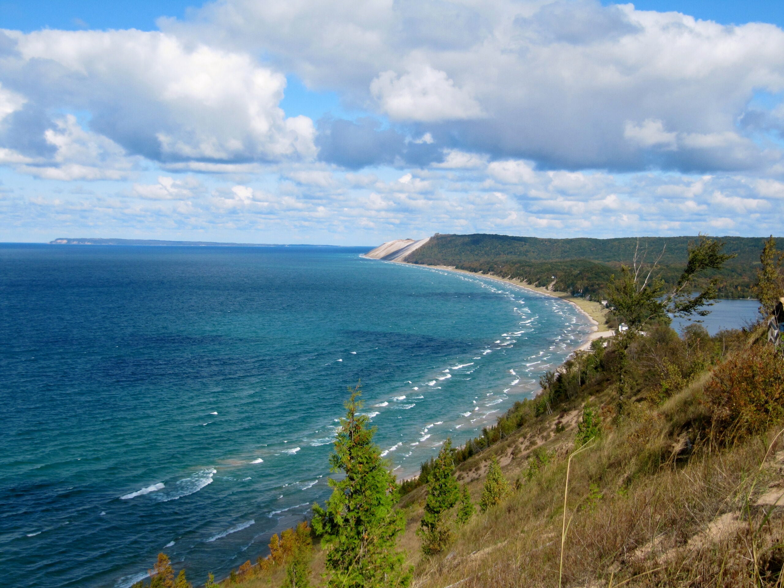 A panoramic view of a curving lakeshore with turquoise water, sandy dunes, and a forested coastline under a partly cloudy blue sky.