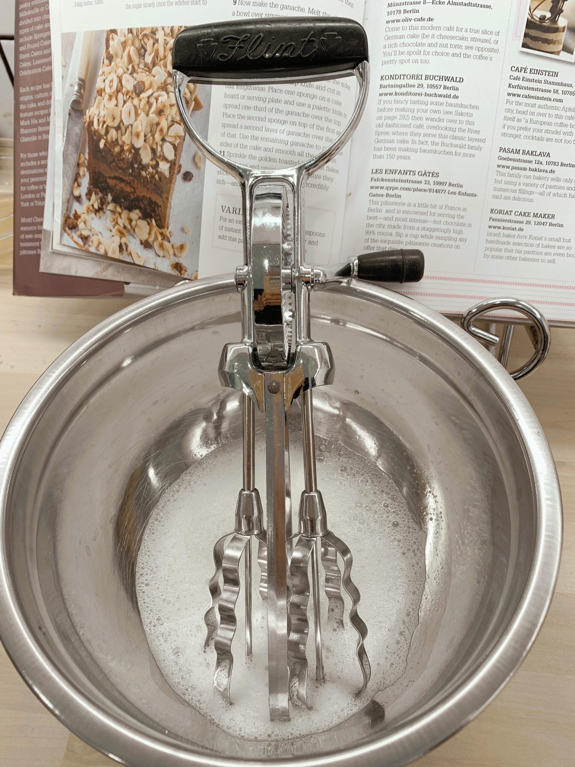 A vintage hand mixer with two metal beaters rests in a stainless steel bowl filled with soapy water, with an open recipe book in the background.
