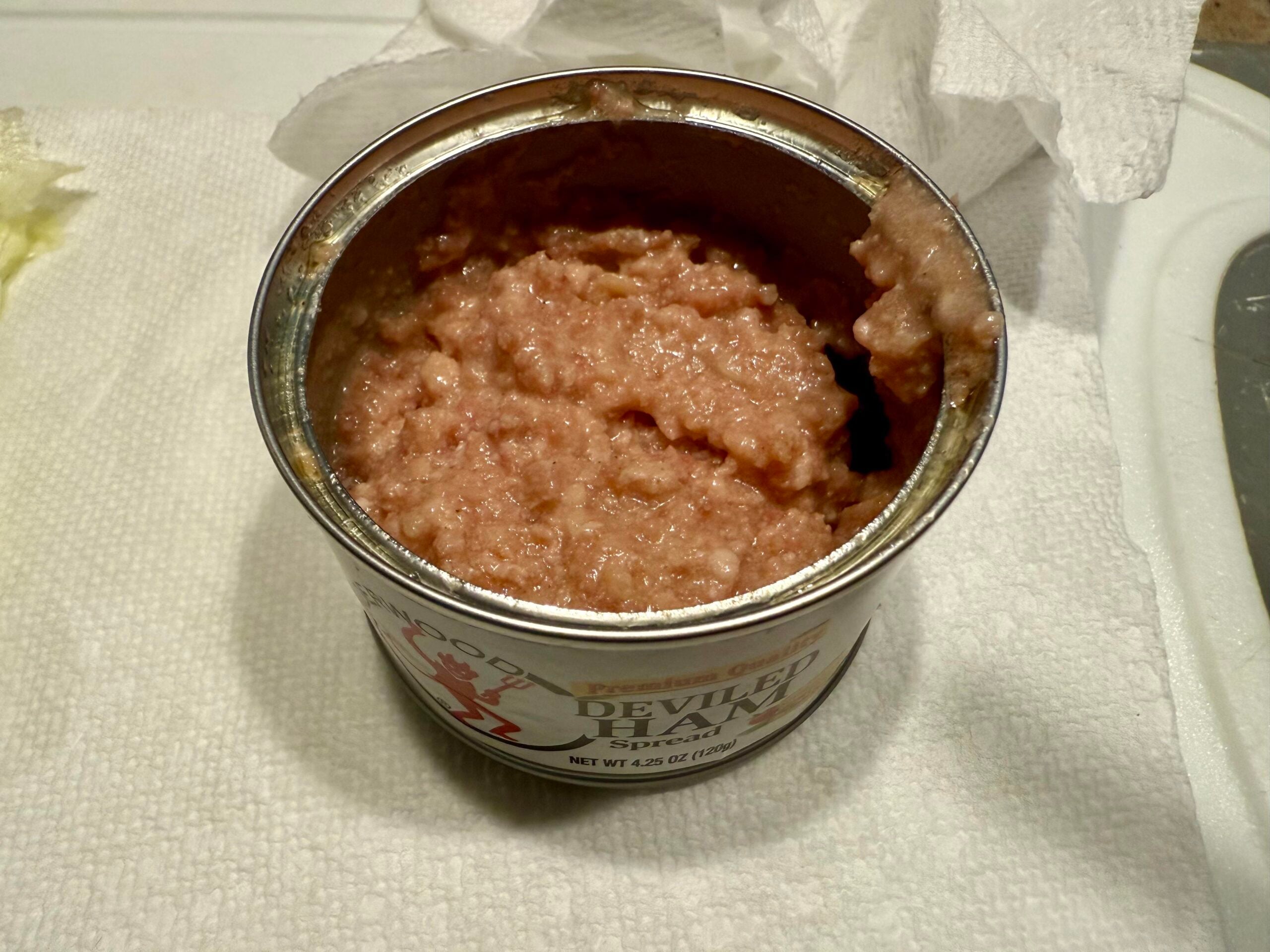 An opened can of deviled ham spread sits on a white paper towel, with its contents showing a soft, pinkish mixture. A crumpled napkin is visible in the background.