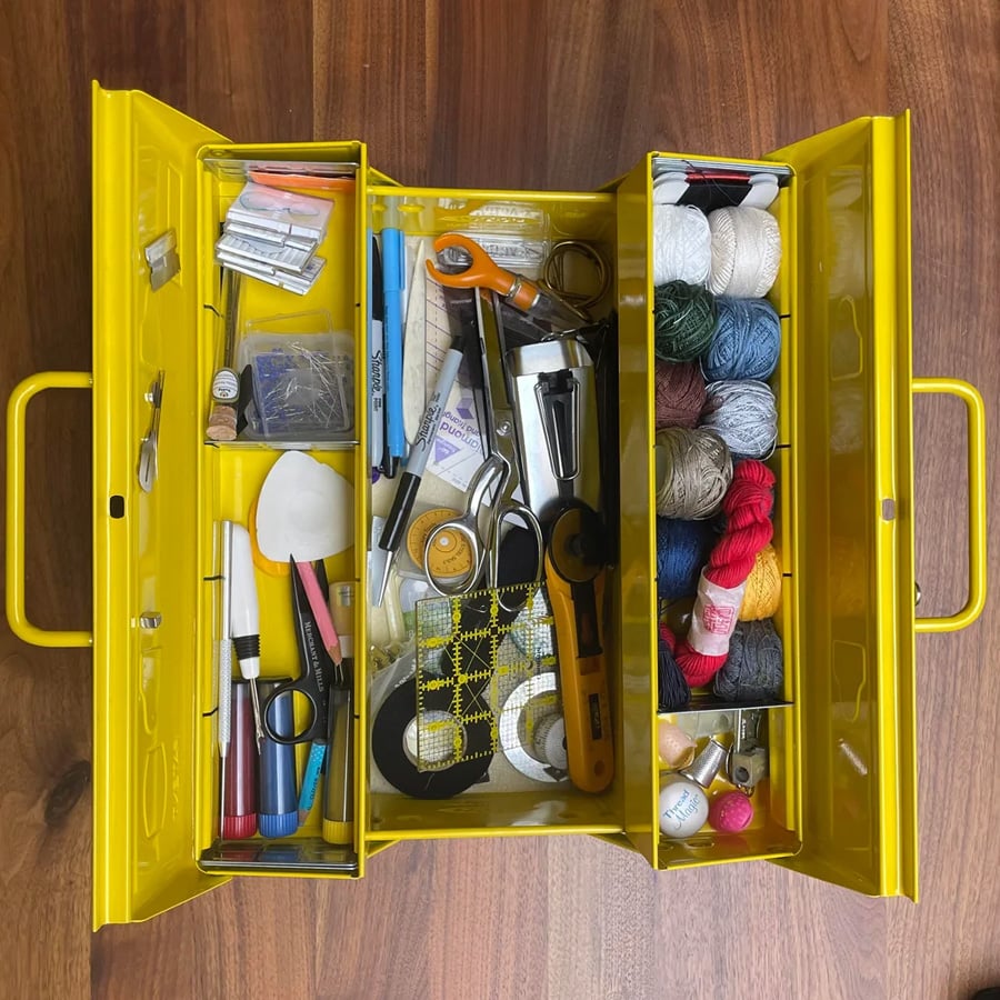 Open yellow sewing box with compartments containing thread spools, scissors, needles, measuring tape, markers, fabric, crochet hooks, and other sewing and crafting supplies on a wooden surface.