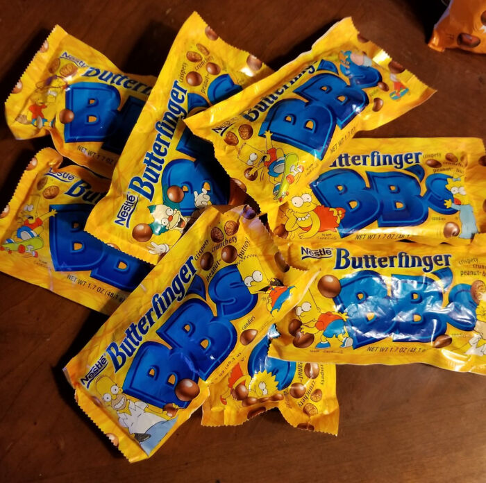 Several yellow packages of Butterfinger BB’s candy featuring The Simpsons characters are scattered on a wooden surface.