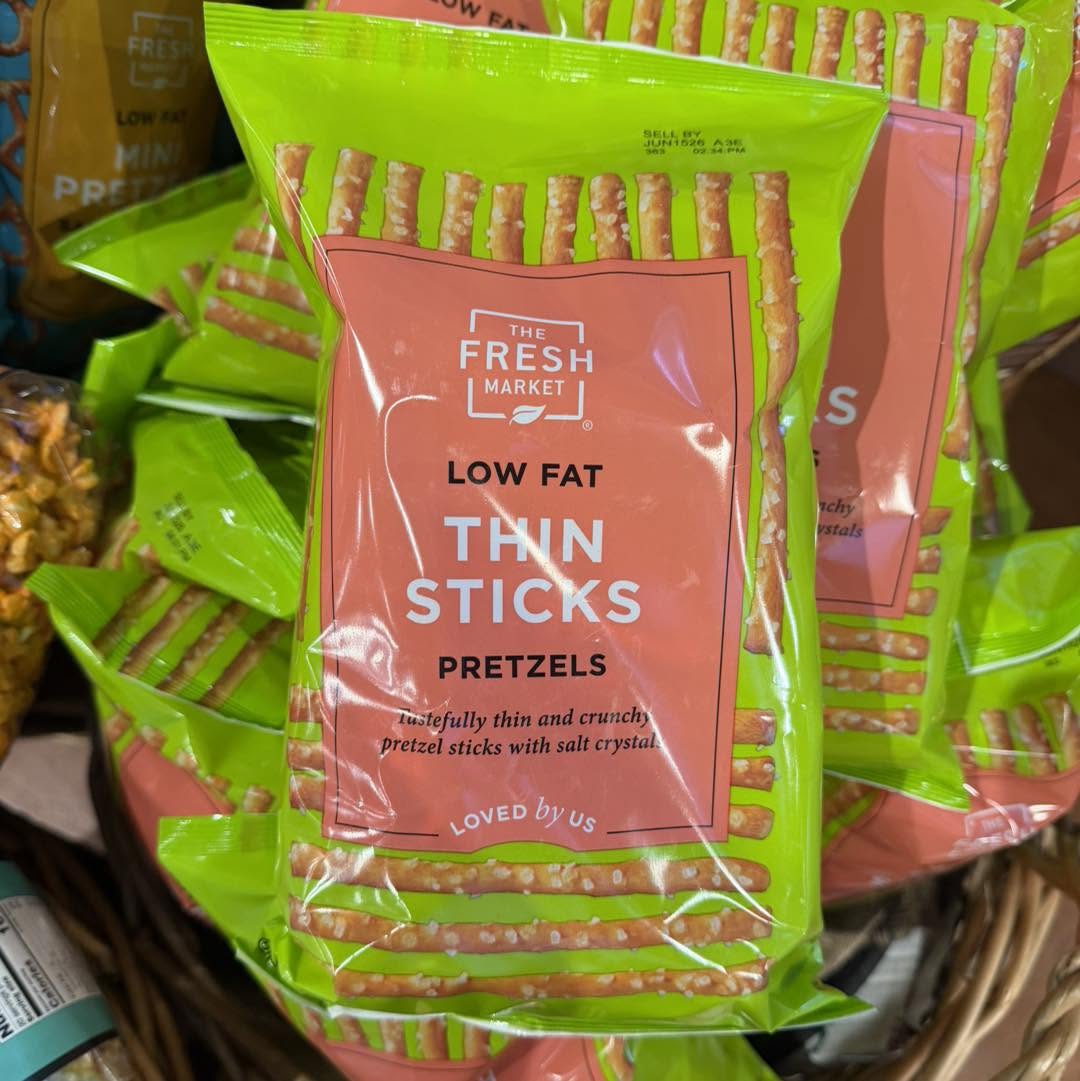 A bag of The Fresh Market Low Fat Thin Sticks Pretzels, featuring green and orange packaging, is displayed among other bags of pretzels.