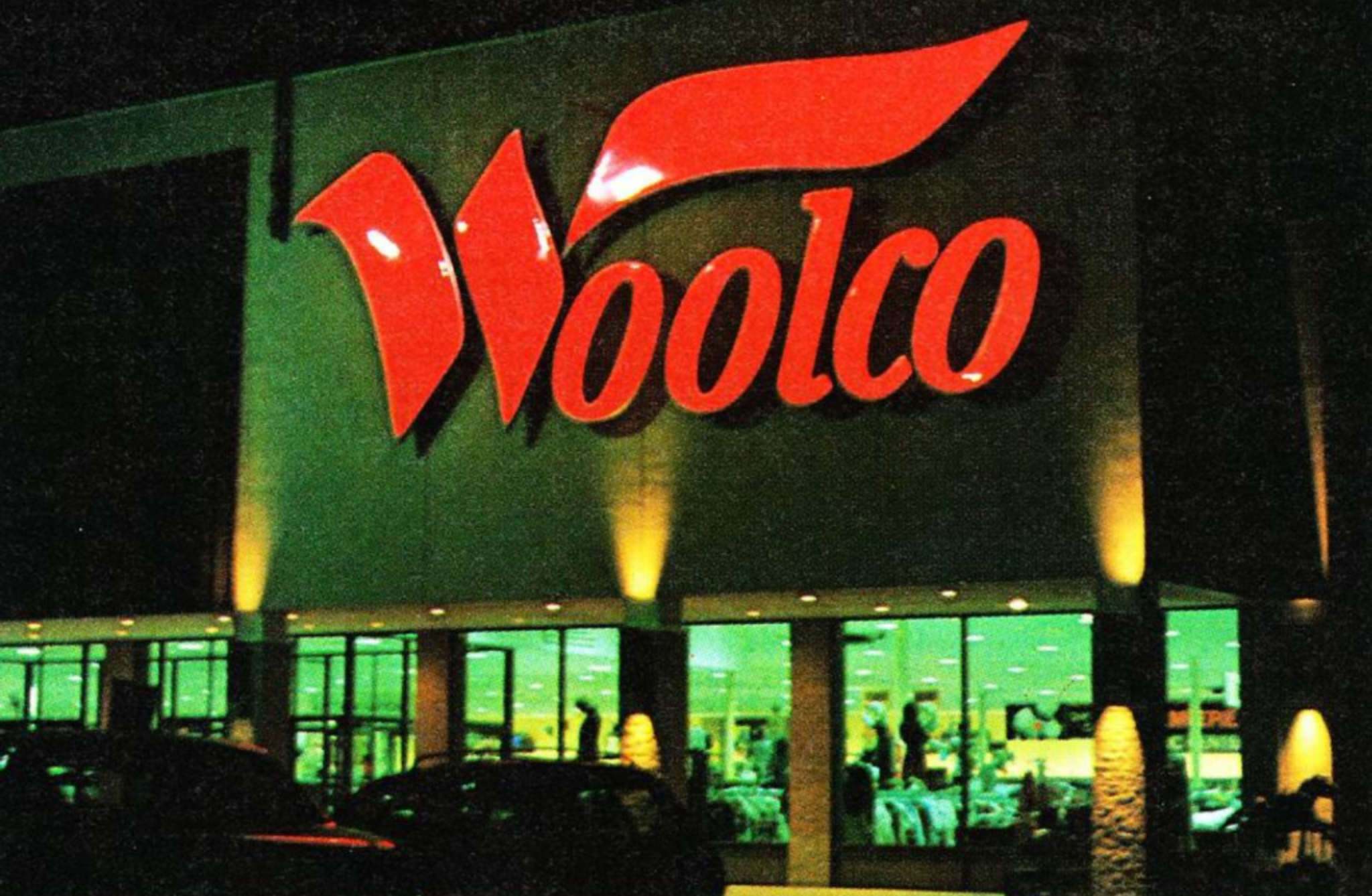 A brightly lit Woolco store at night, featuring a large red Woolco sign above the entrance and large glass windows showcasing merchandise inside.