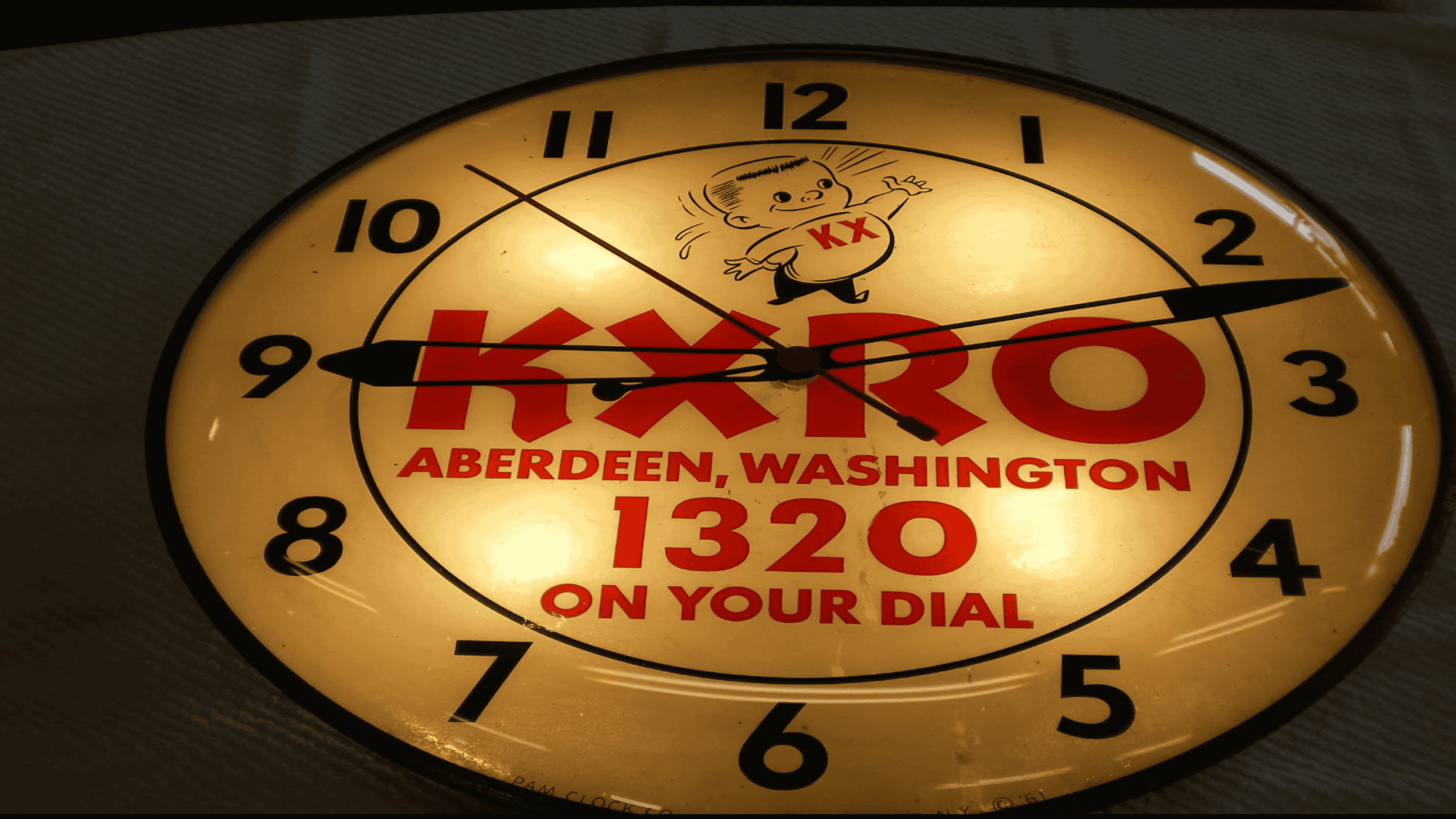 A round clock with “KXRO Aberdeen, Washington 1320 on your dial” printed in red and a cartoon baby above the text. The hands show 10:10. The clock is warmly lit.