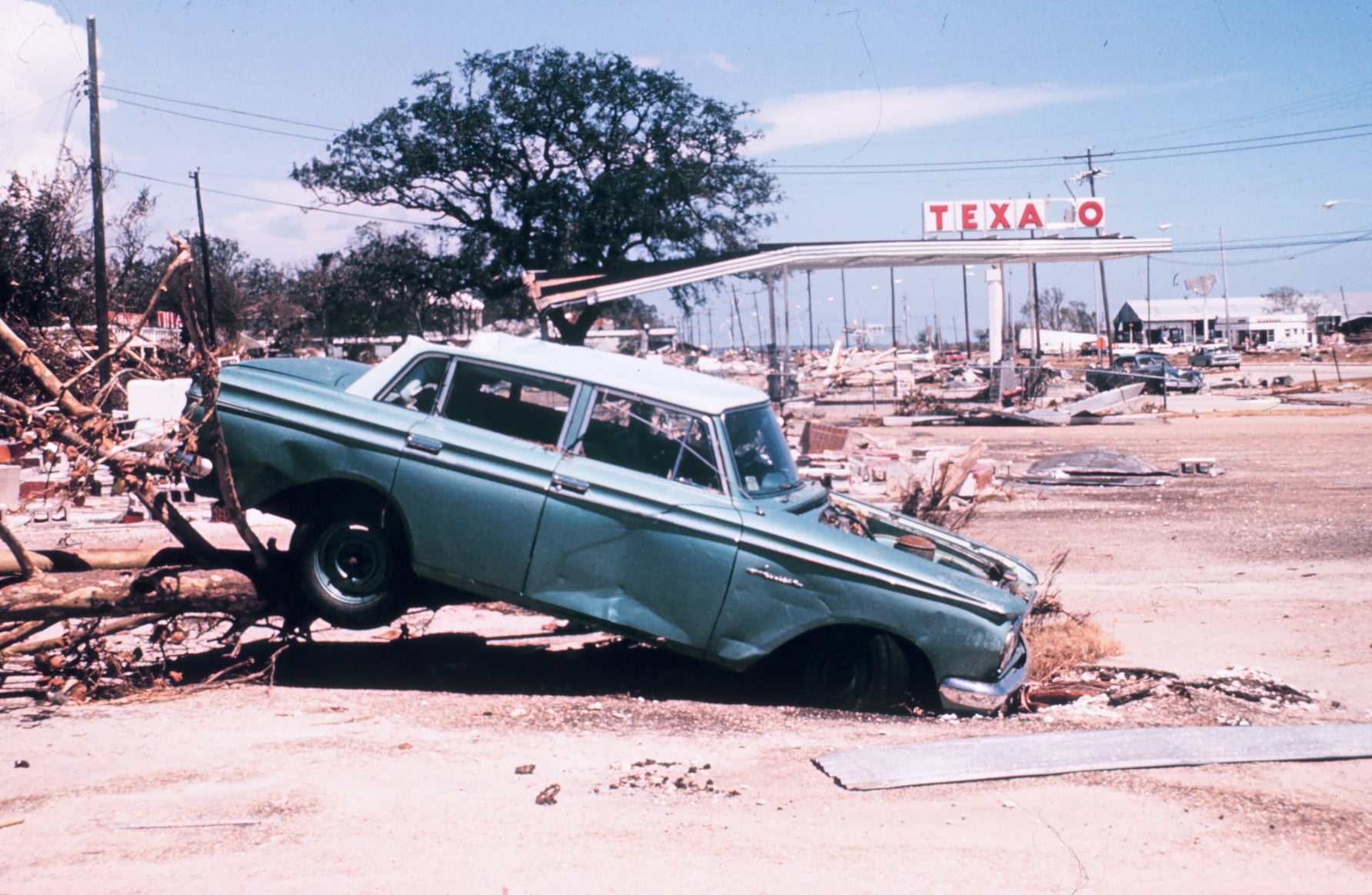 A battered green car is half-sunken in the ground amid storm debris. In the background, a damaged Texaco gas station and scattered wreckage are visible under a blue sky.