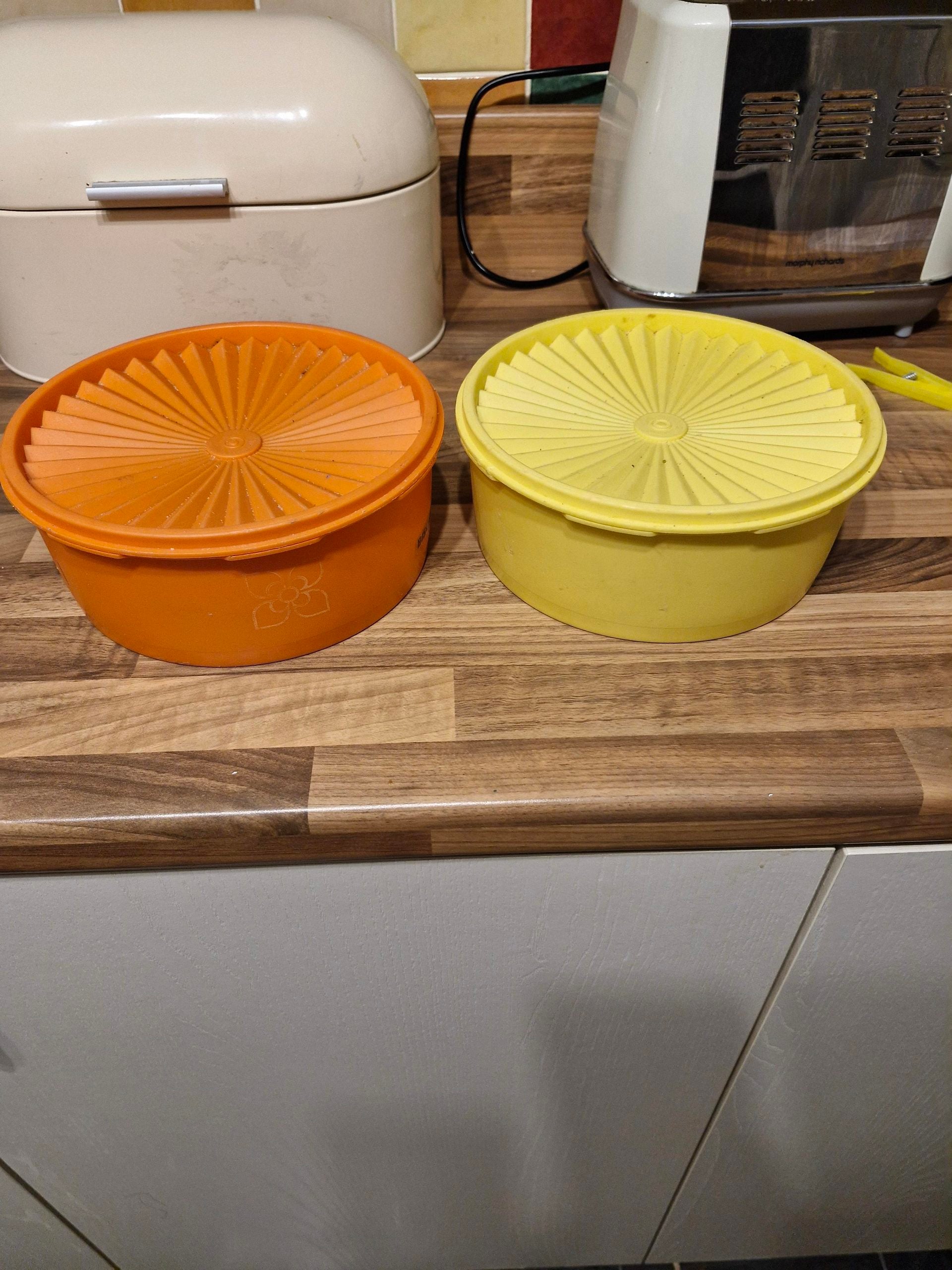 Two round plastic containers, one orange and one yellow, with matching lids, sit side by side on a wooden kitchen counter. Kitchen appliances are visible in the background.