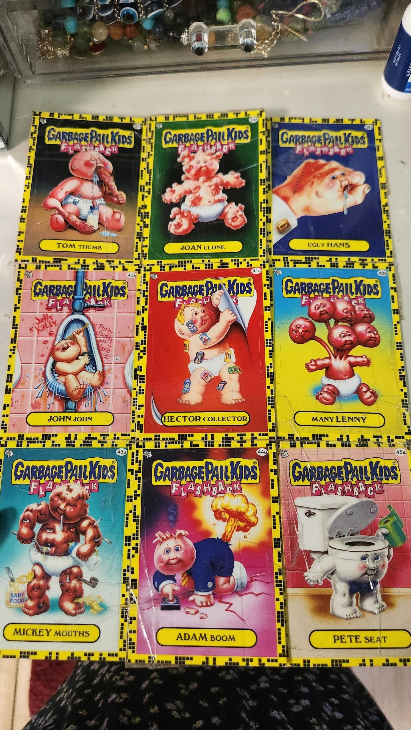 Nine Garbage Pail Kids cards laid out in three rows on a surface. Each card features a unique cartoon character with a humorous, pun-filled name and colorful artwork. Jewelry is visible in the background.
