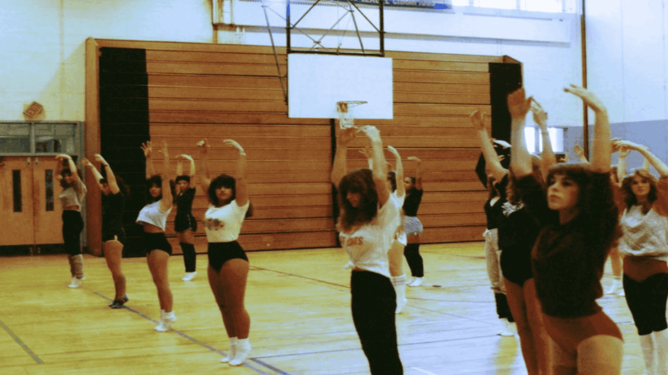A group of women in leotards and gym clothes practice dance or cheerleading moves in a school gymnasium, arms raised, under a basketball hoop.