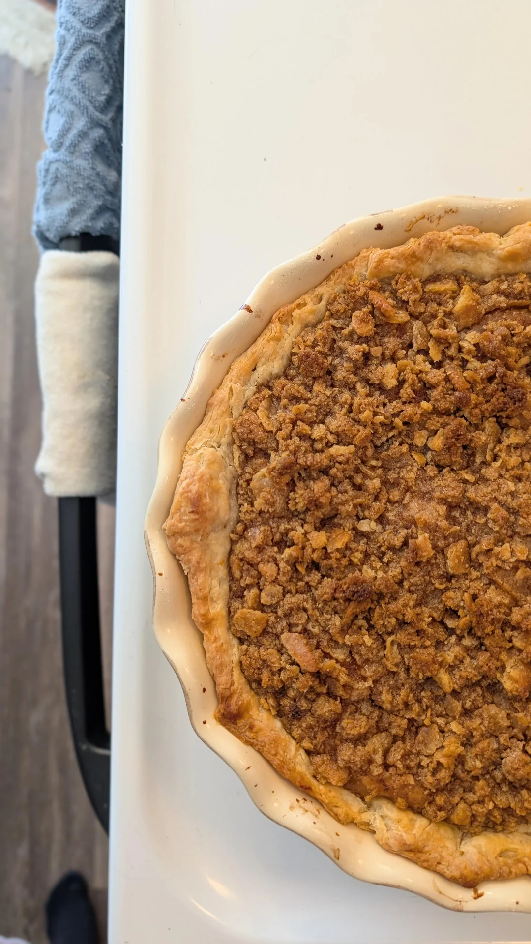 A baked pie with a crumbly brown topping sits in a cream-colored dish on a white surface next to a blue oven mitt.