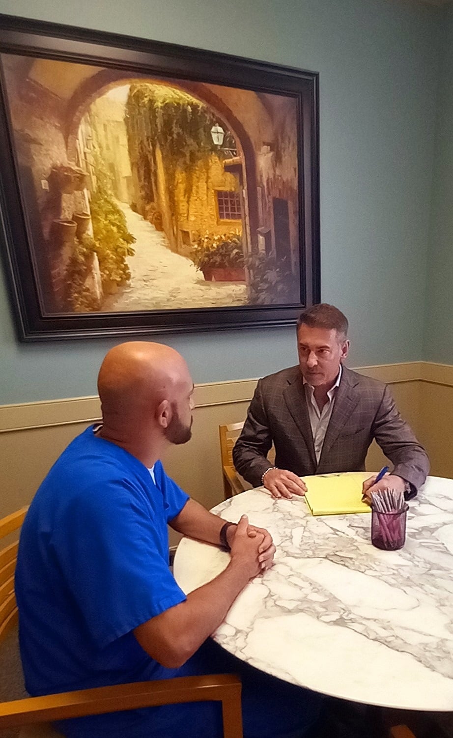 Two men sit at a round marble table having a discussion. One man wears blue scrubs, and the other wears a gray suit, holding a yellow notepad. A painting of a European street hangs on the wall behind them.