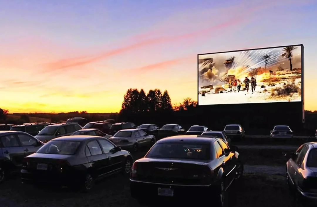 Cars parked at a drive-in movie theater at sunset, with an action scene featuring explosions and soldiers displayed on the large outdoor screen.