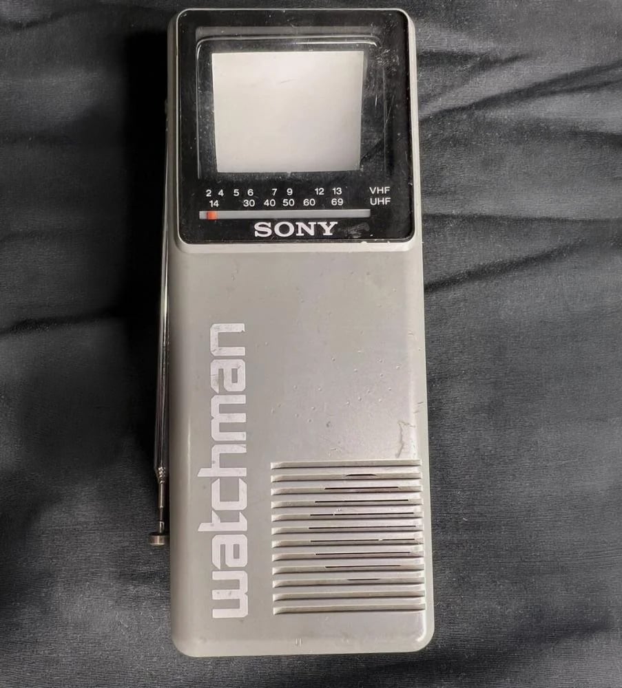 A vintage Sony Watchman portable TV with a small screen, numeric channel markers, and a silver body lying on a dark surface. The word “watchman” is printed vertically on the front.